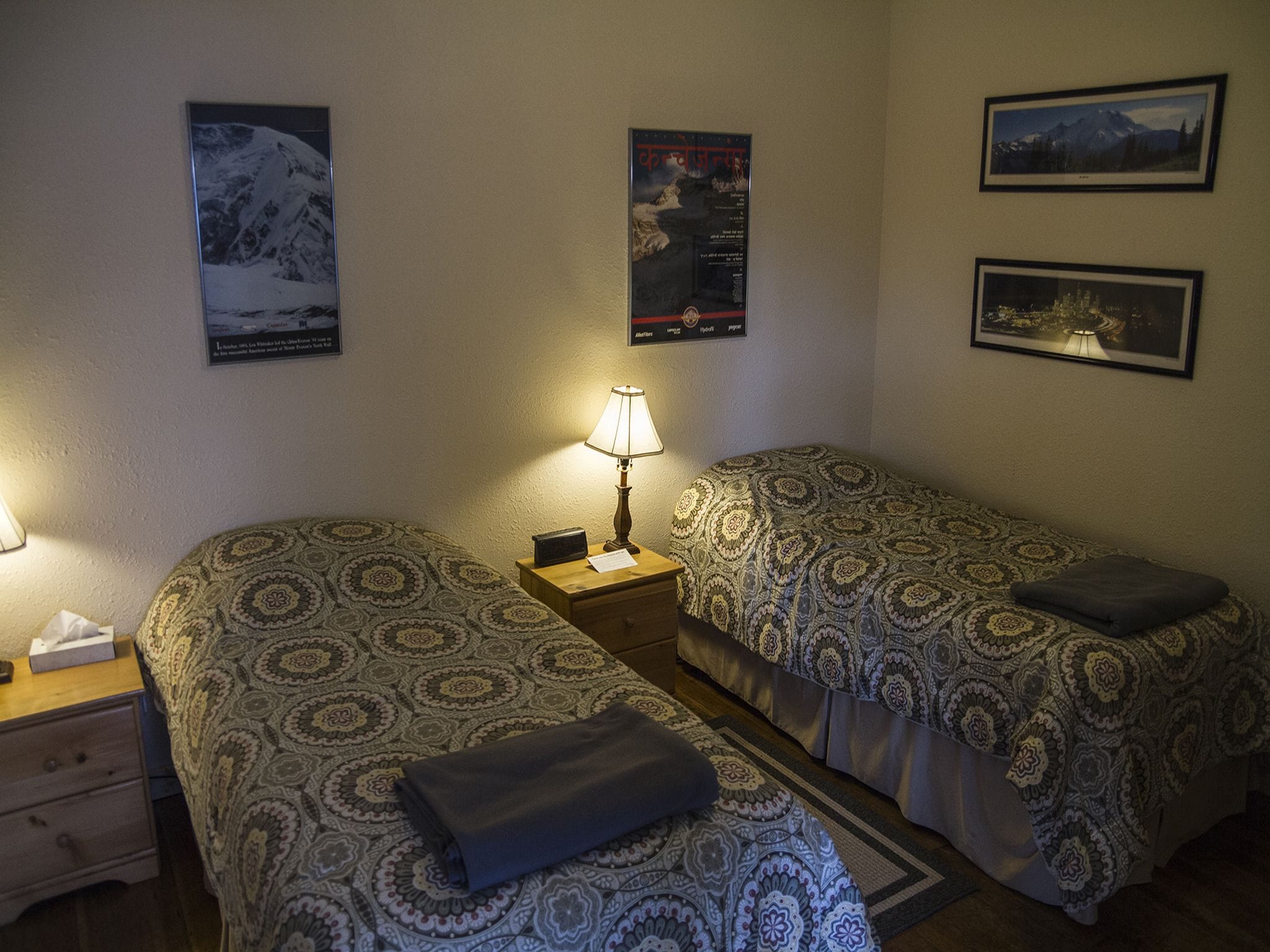 Habitación de hotel con dos camas, una mesita de noche con lámpara y tres cuadros enmarcados en la pared.