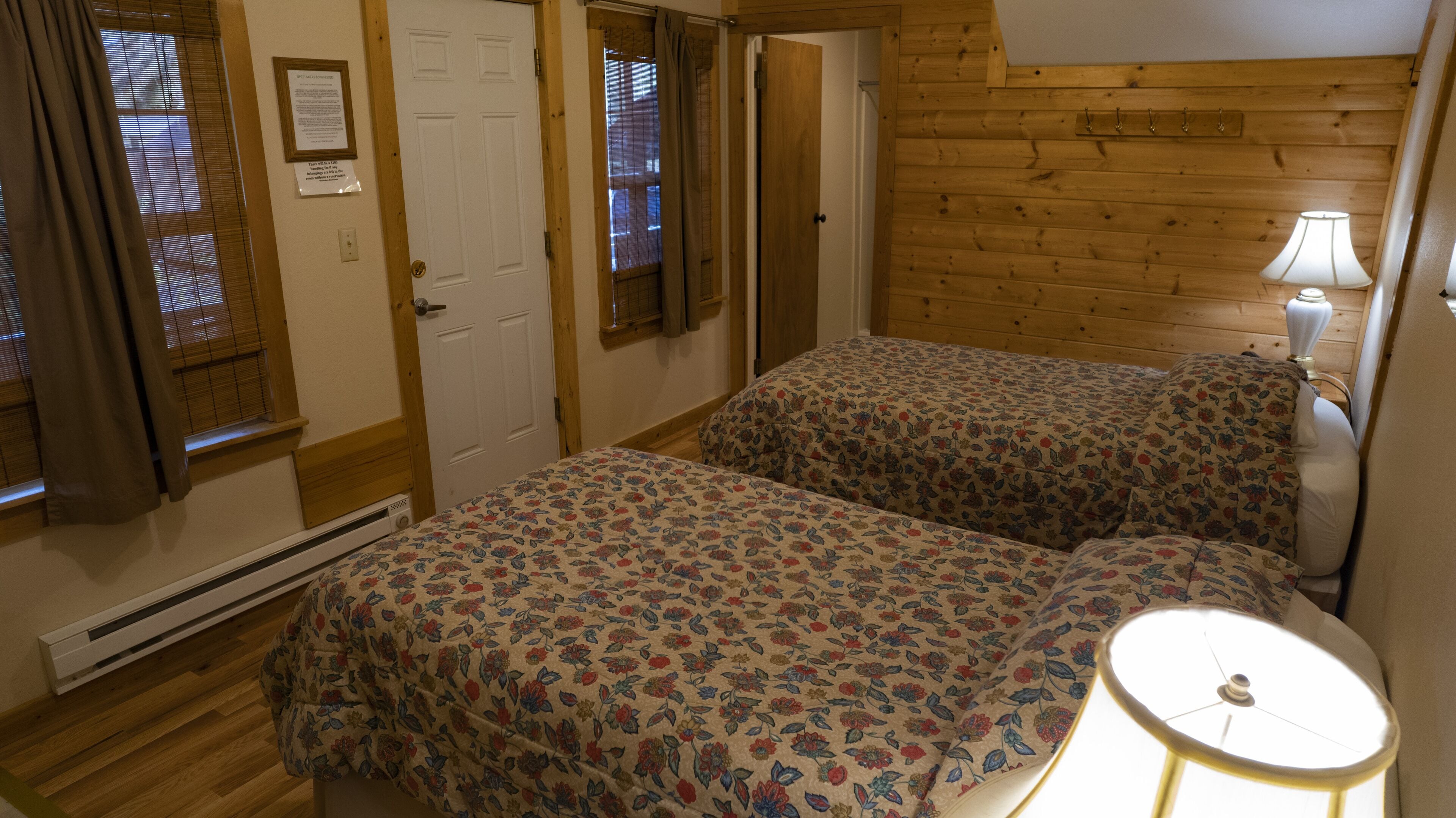 A room with two beds, wooden walls, a heater, a lamp, and a door with a sign.