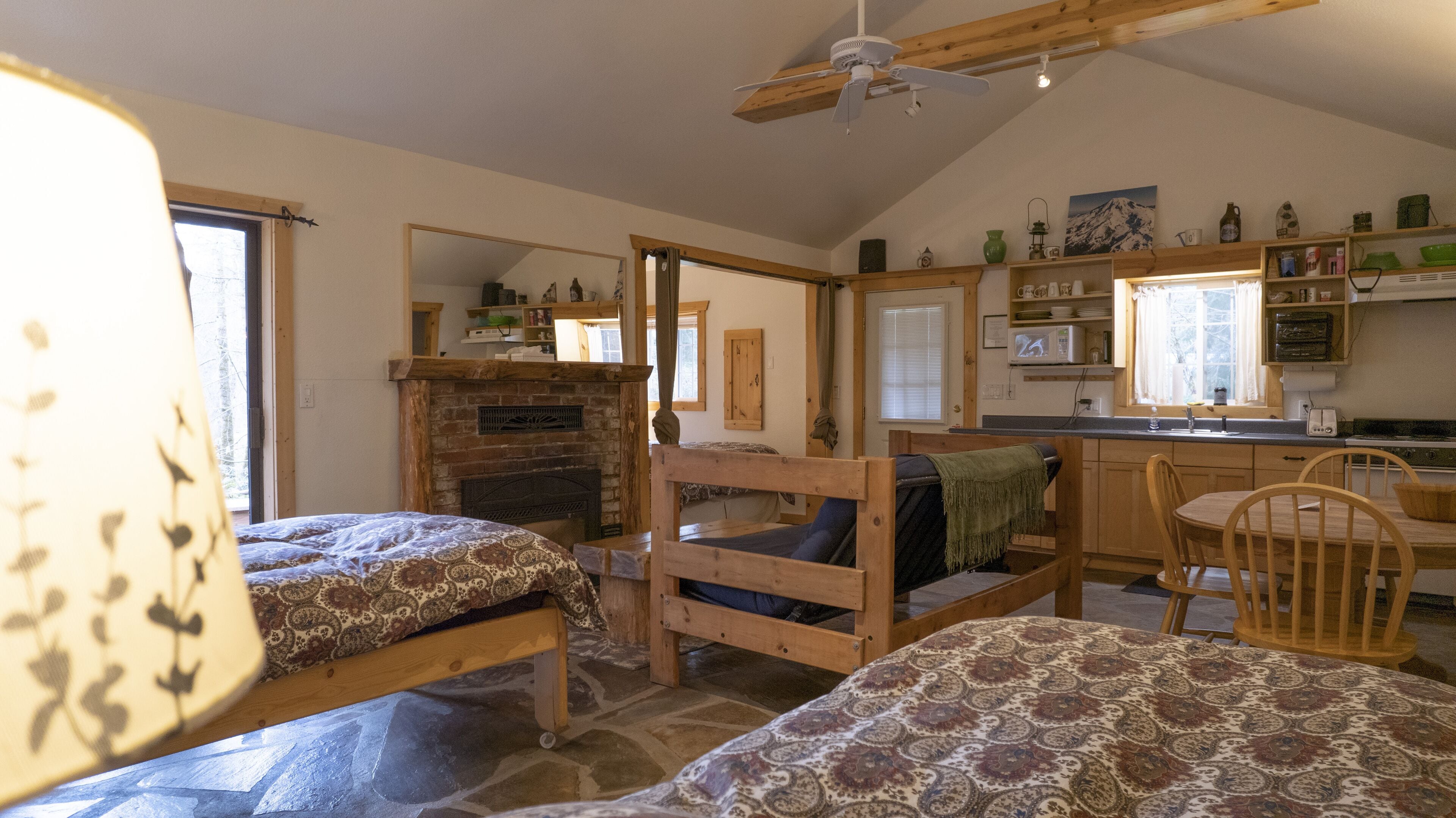 A cozy room with two wooden beds, a fireplace, a dining table with chairs, and a kitchen area with shelves and a window.