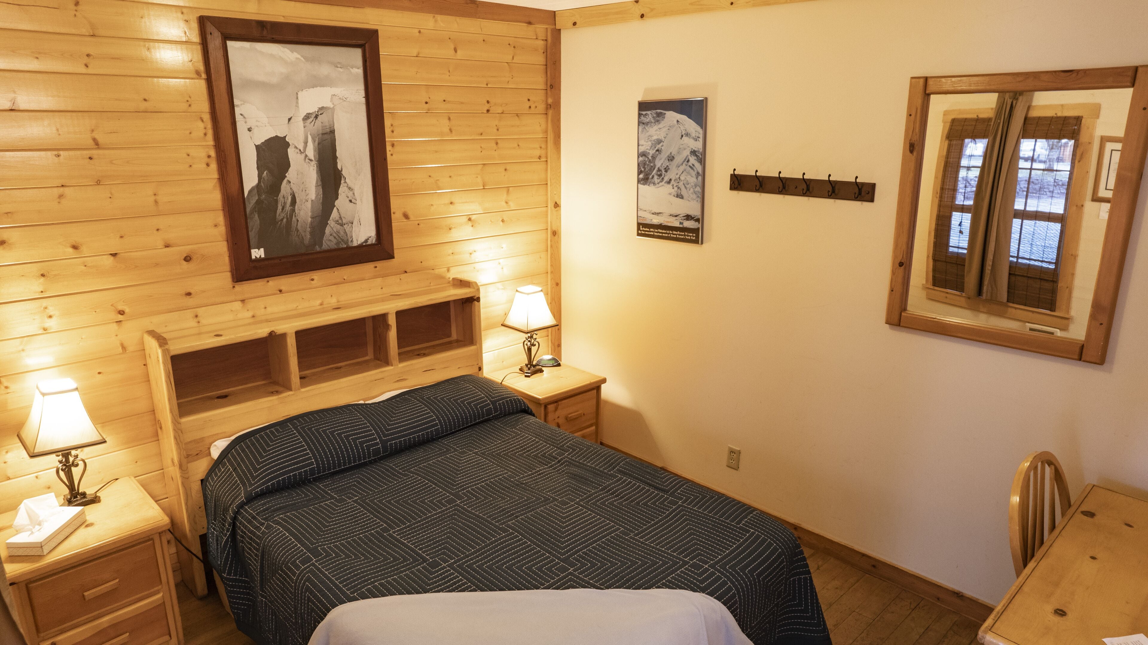 A wooden-paneled room with a bed, two bedside tables with lamps, a framed picture on the wall, a window with curtains, and a small wooden chair.