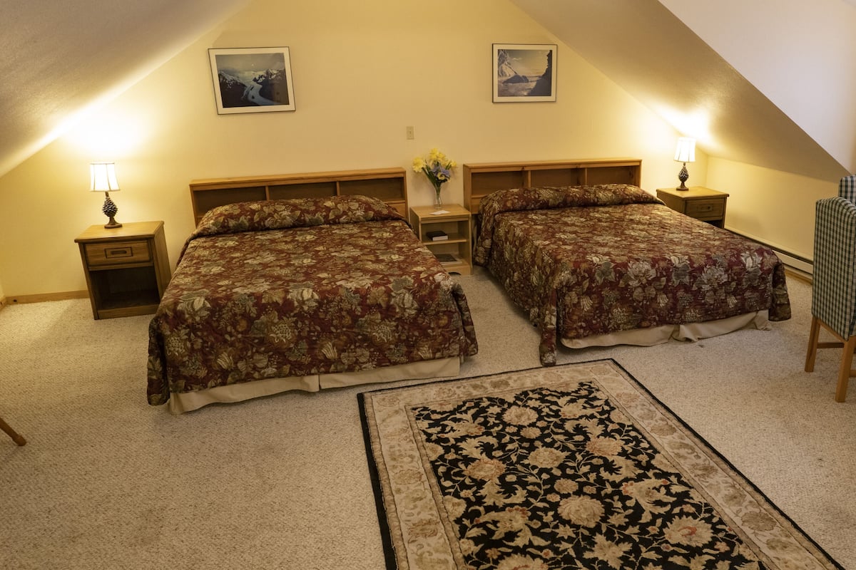 Ein gemütliches Zimmer mit zwei Betten, einem Schreibtisch und einem Stuhl. Zwei Nachttische mit Lampen, ein kleiner Tisch und ein Stuhl sind ebenfalls vorhanden.