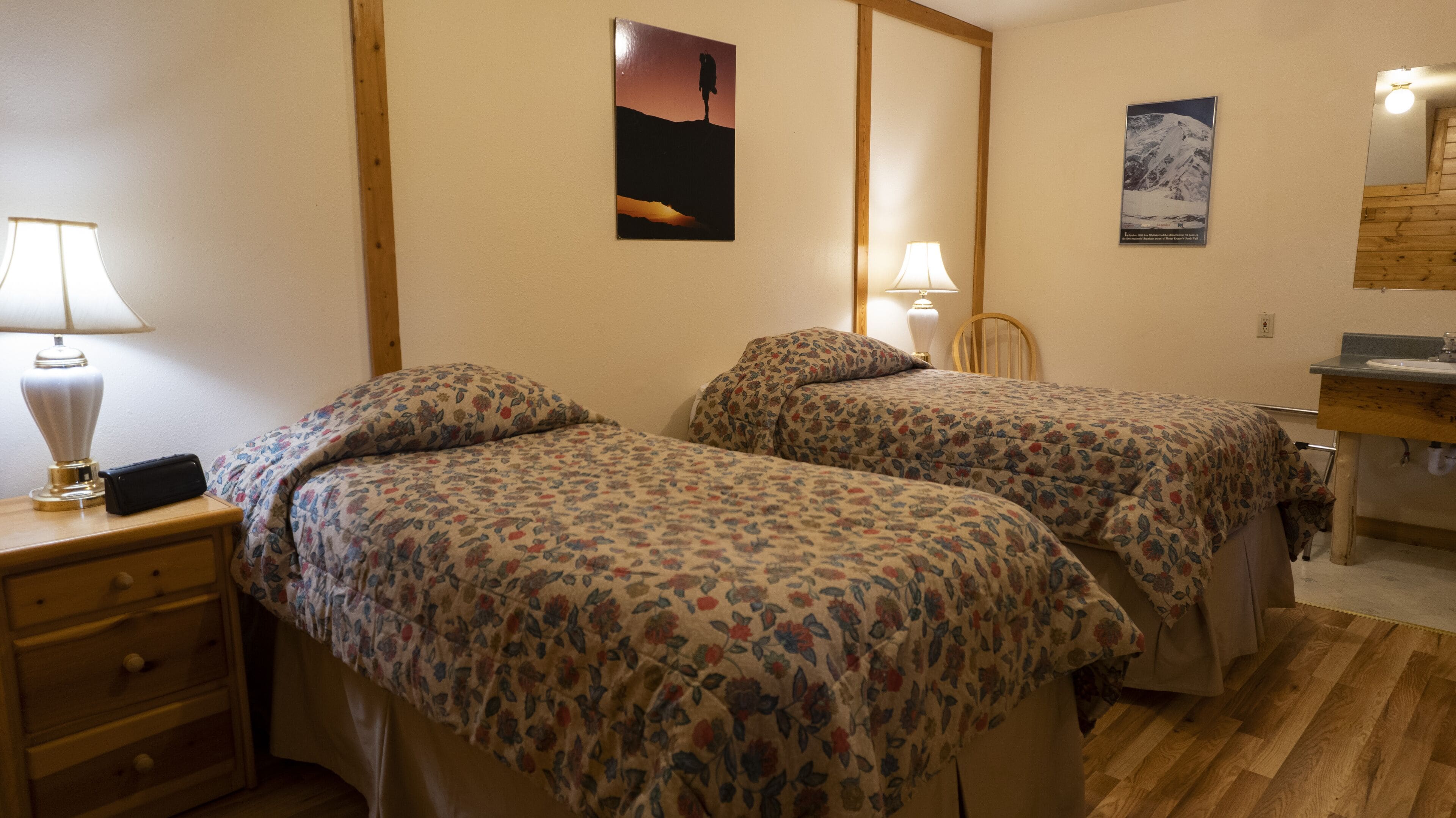 A room with two beds, a wooden nightstand with a lamp, a chair, a wooden desk with a lamp, and a bathroom with a sink and mirror.