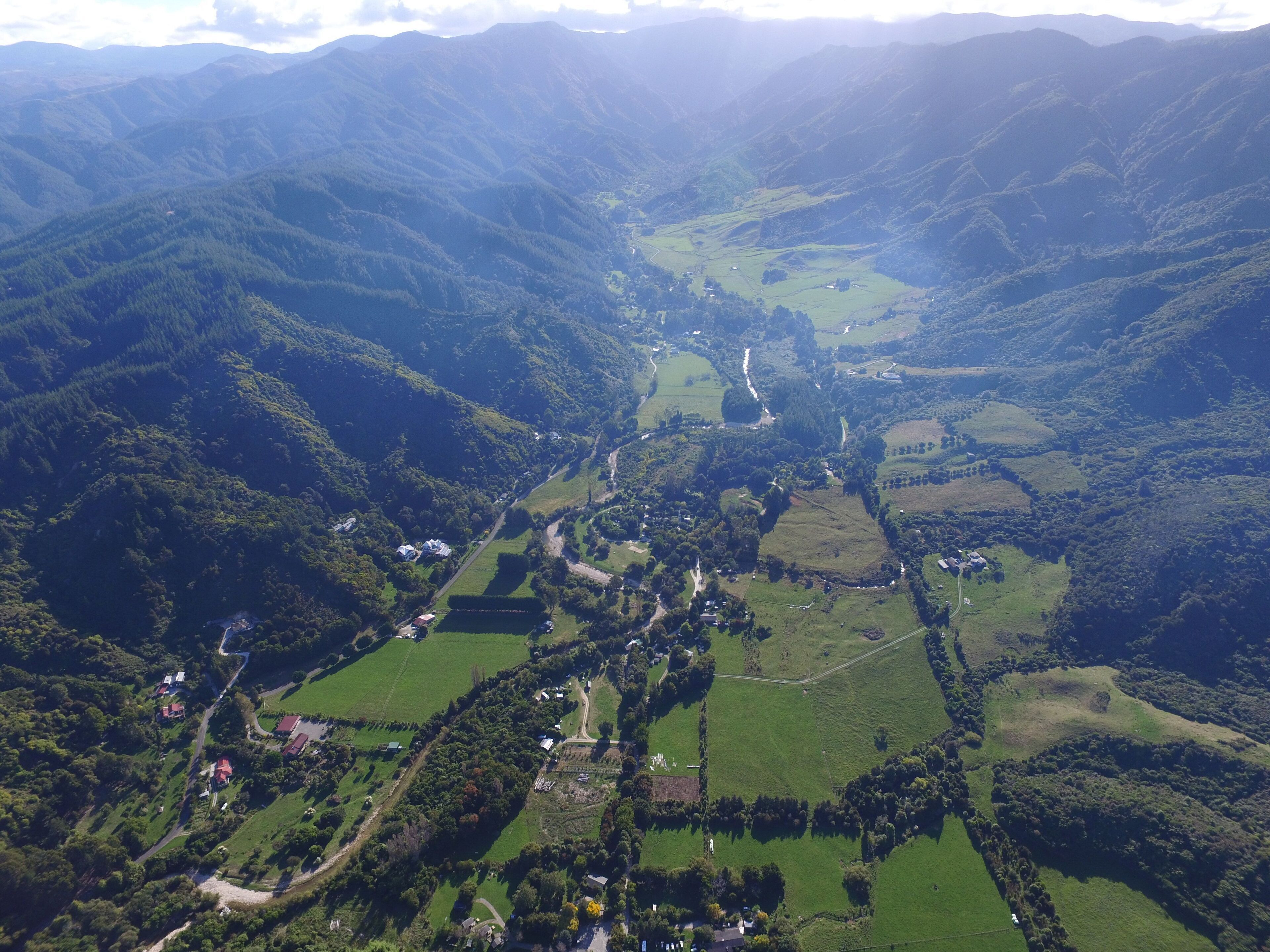 Luchtbeeld van een vallei met groene velden, verspreide huizen en een kronkelende weg.