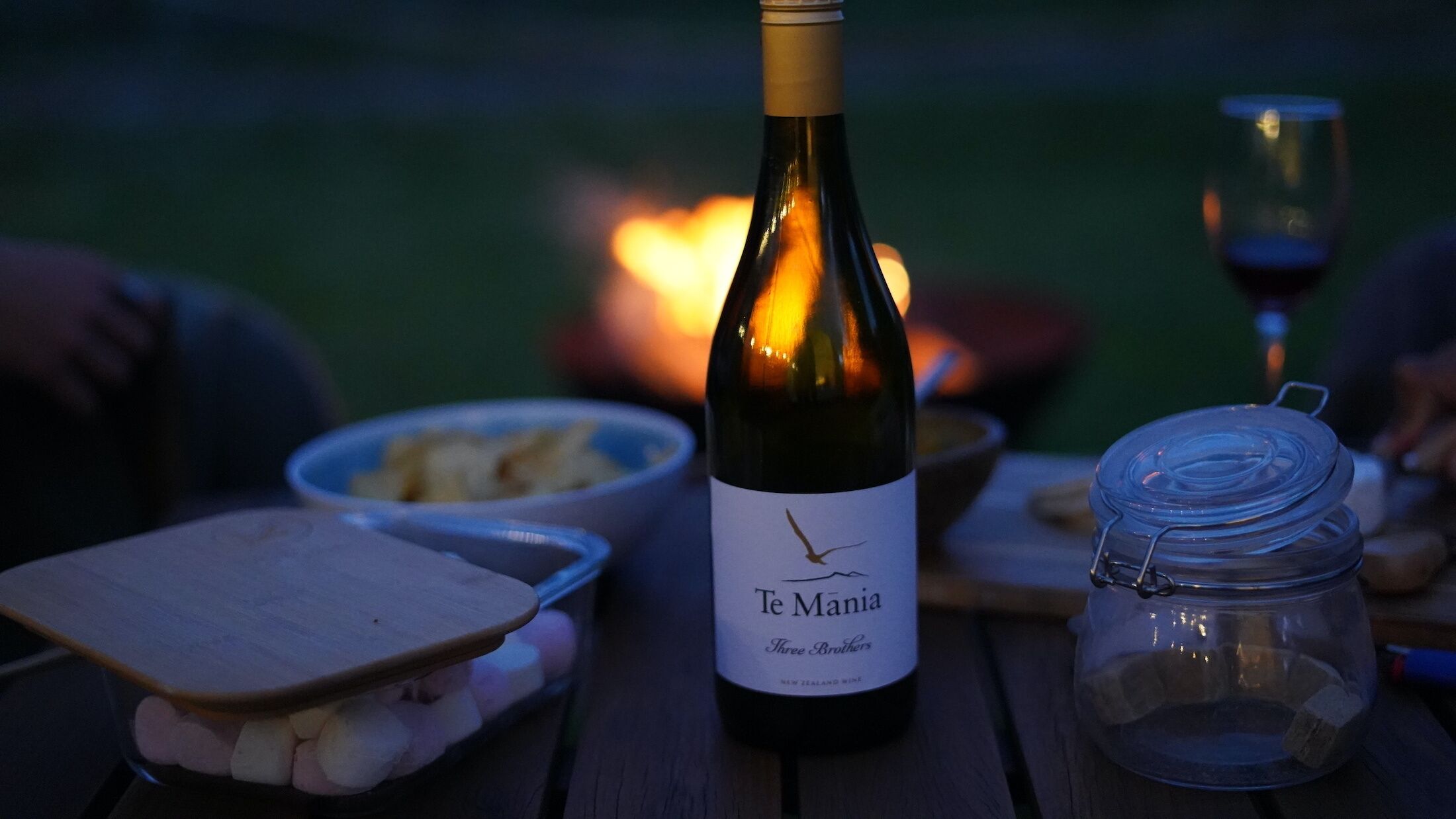 A bottle of Te Manoa wine, a wooden cutting board with food, a glass jar, and a bowl of chips on a table.