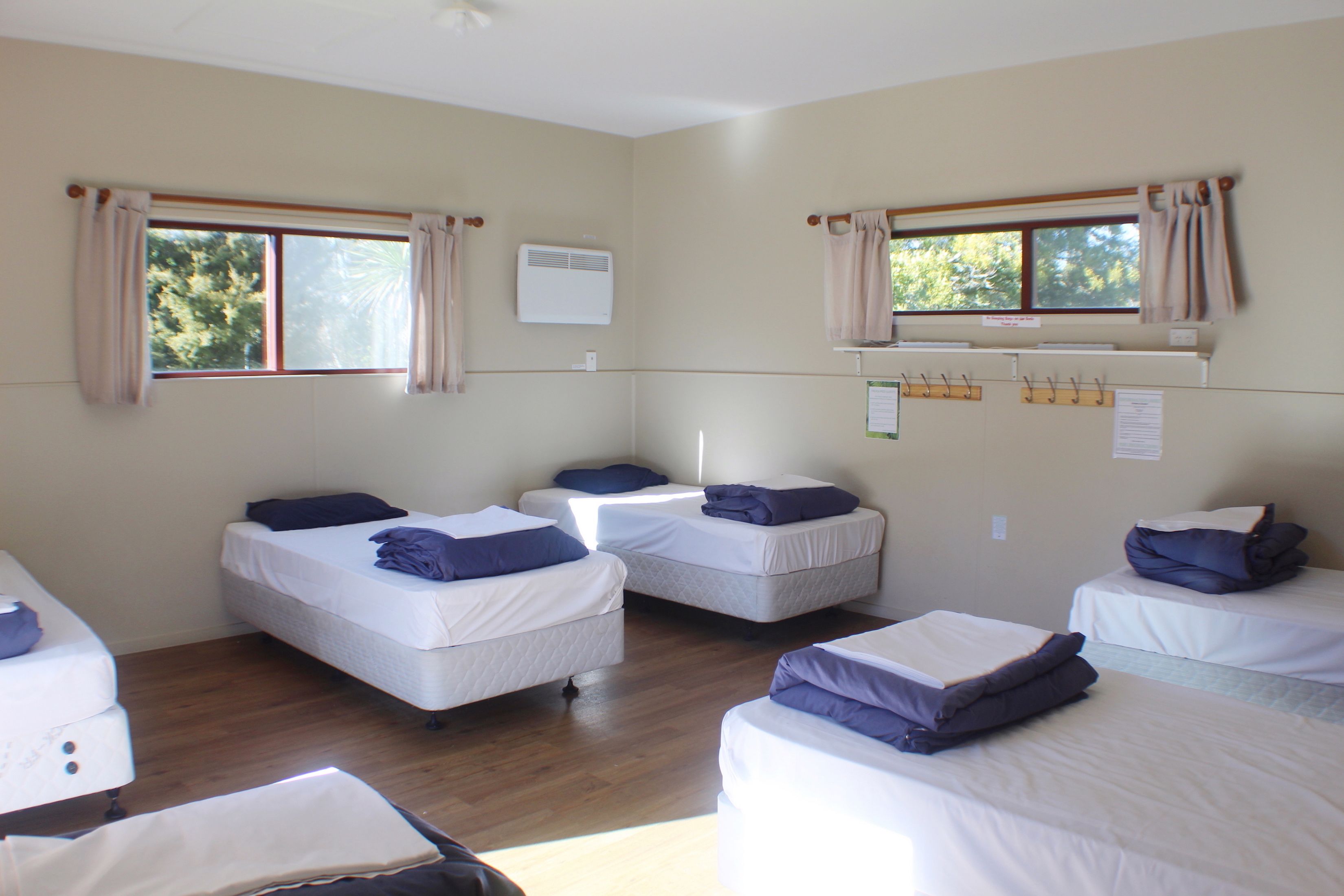 Una habitación con cuatro camas individuales, cada una con almohada y manta, suelo de madera y ventanas con cortinas.