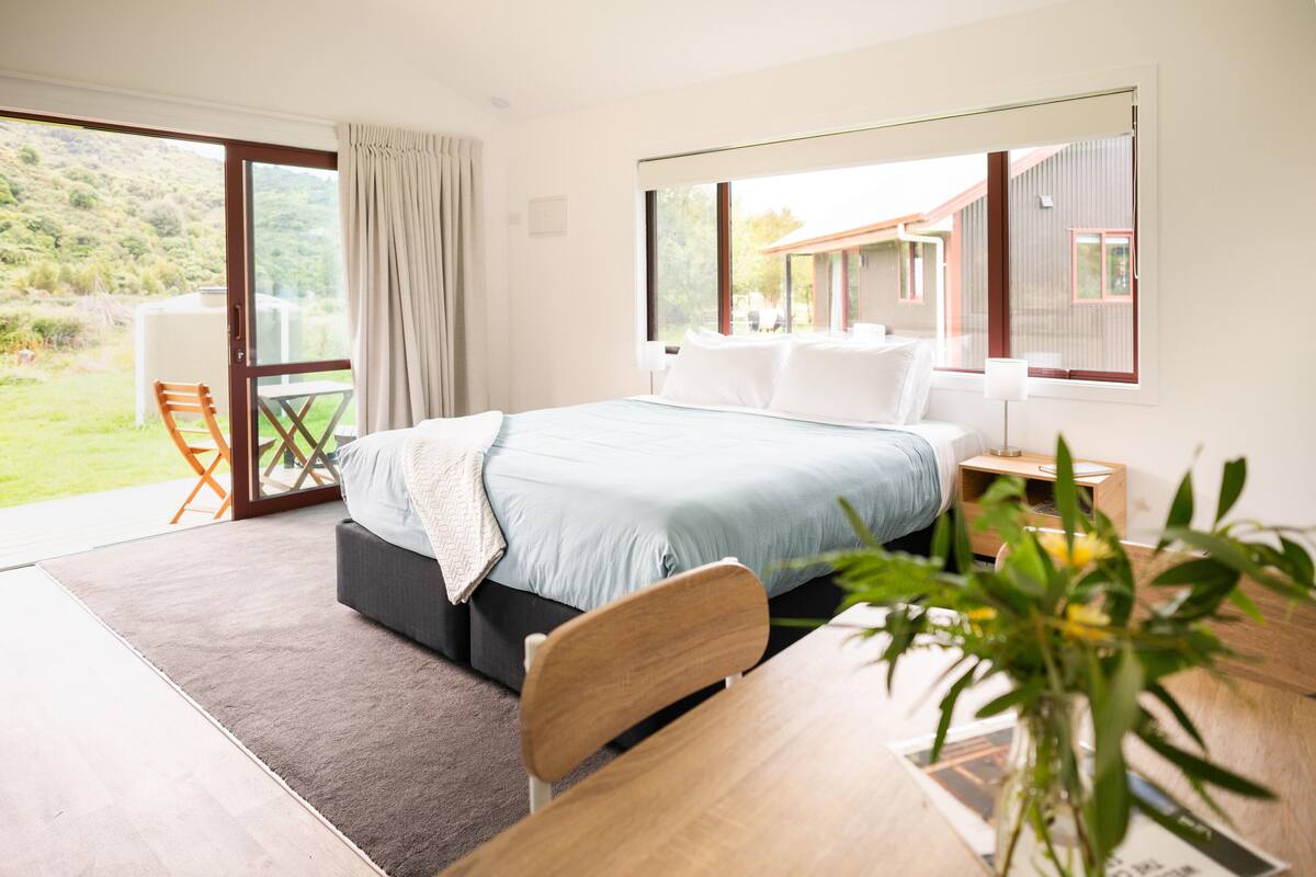 A modern bedroom with a large bed, a wooden nightstand, a desk, and a chair. There is a sliding glass door leading to a balcony with a chair and a table.