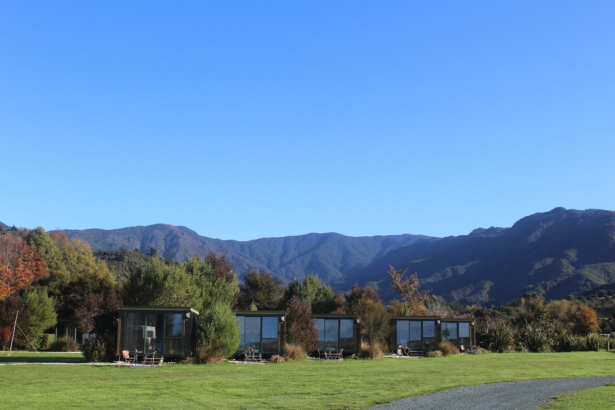 A modern building with large glass windows, surrounded by a grassy field and trees, with mountains in the background.
