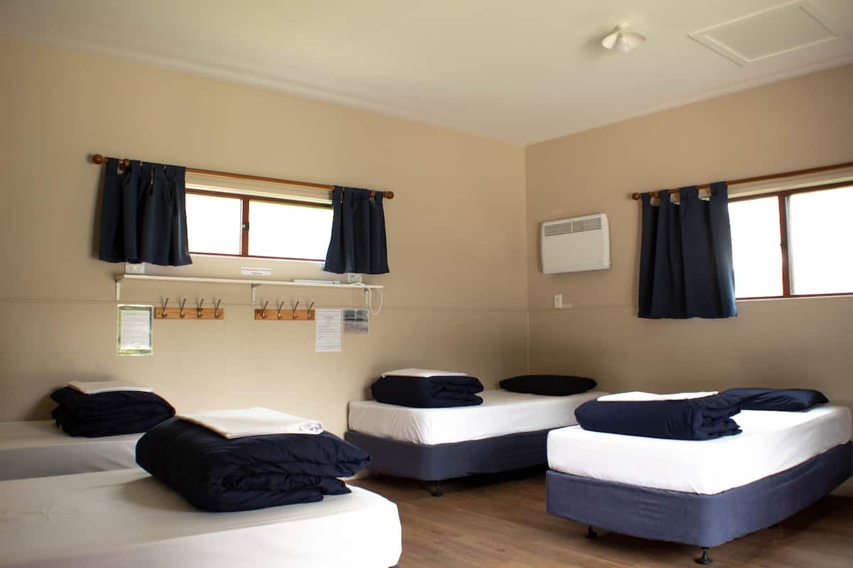 A hotel room with four single beds, each with a dark blue blanket and pillow. There are two windows with dark blue curtains, and an air conditioning unit mounted on the wall.