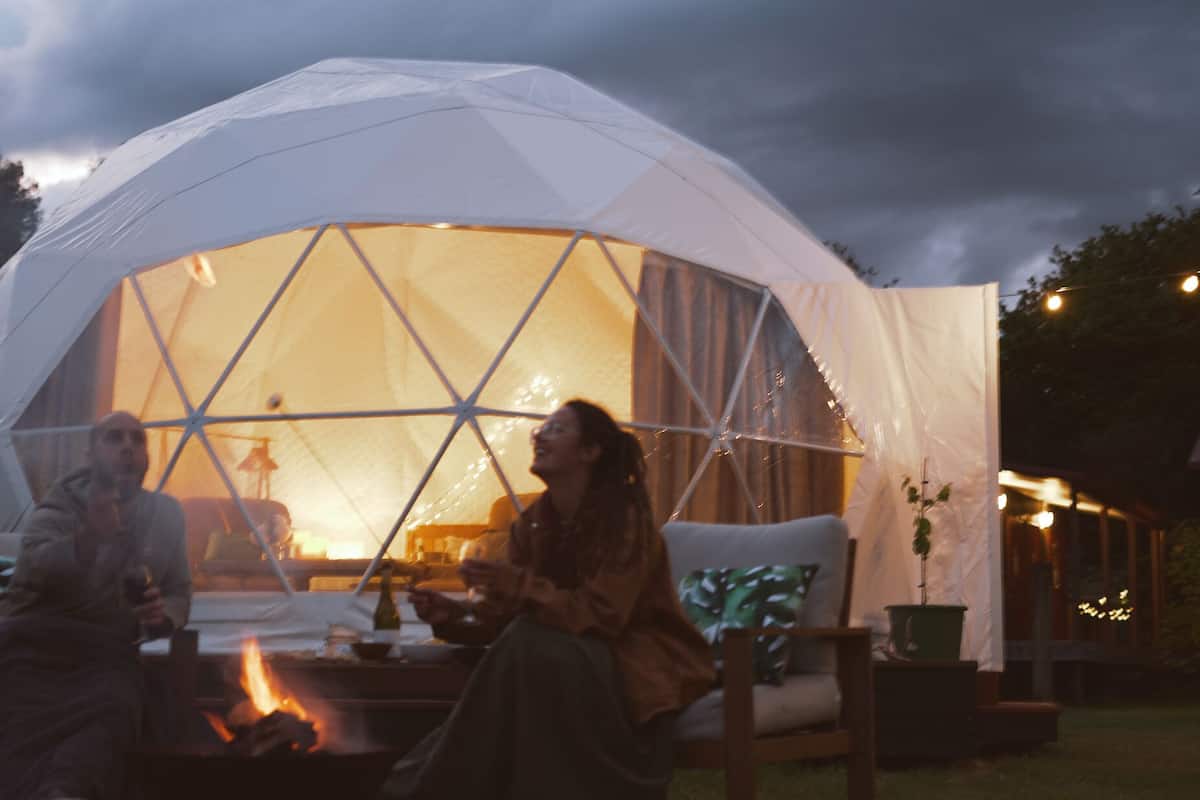 A geodesic dome tent with a fire in the center, surrounded by a garden setting.