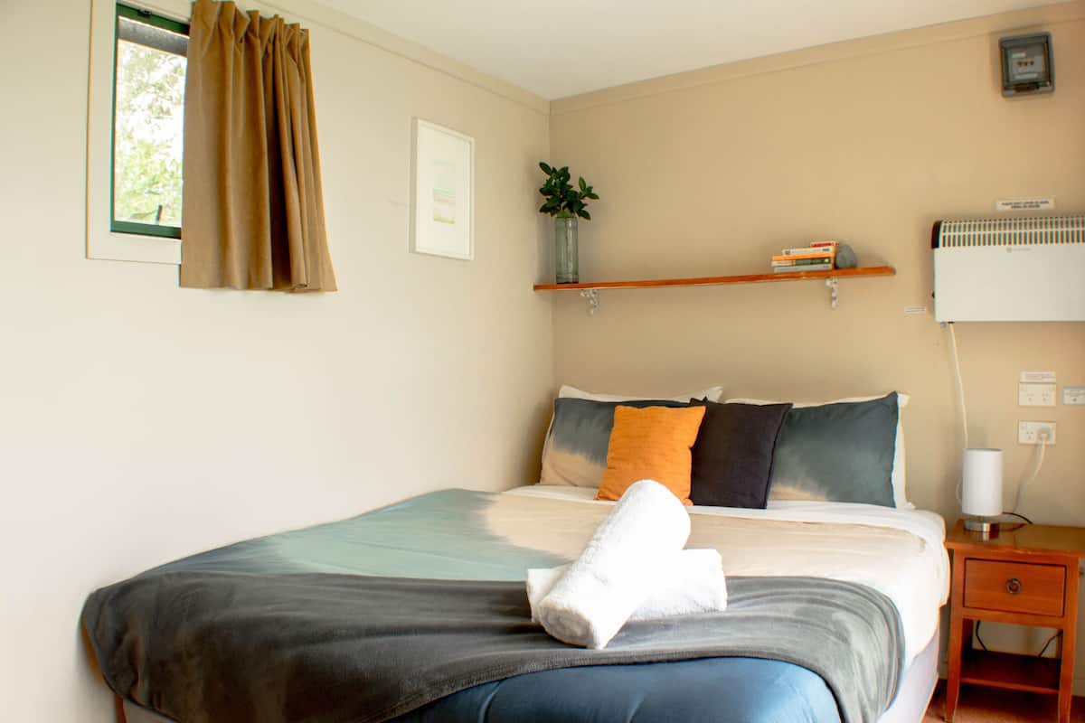 A neatly made bed with a rolled towel, a bedside table with a lamp, and a shelf with books and a plant.