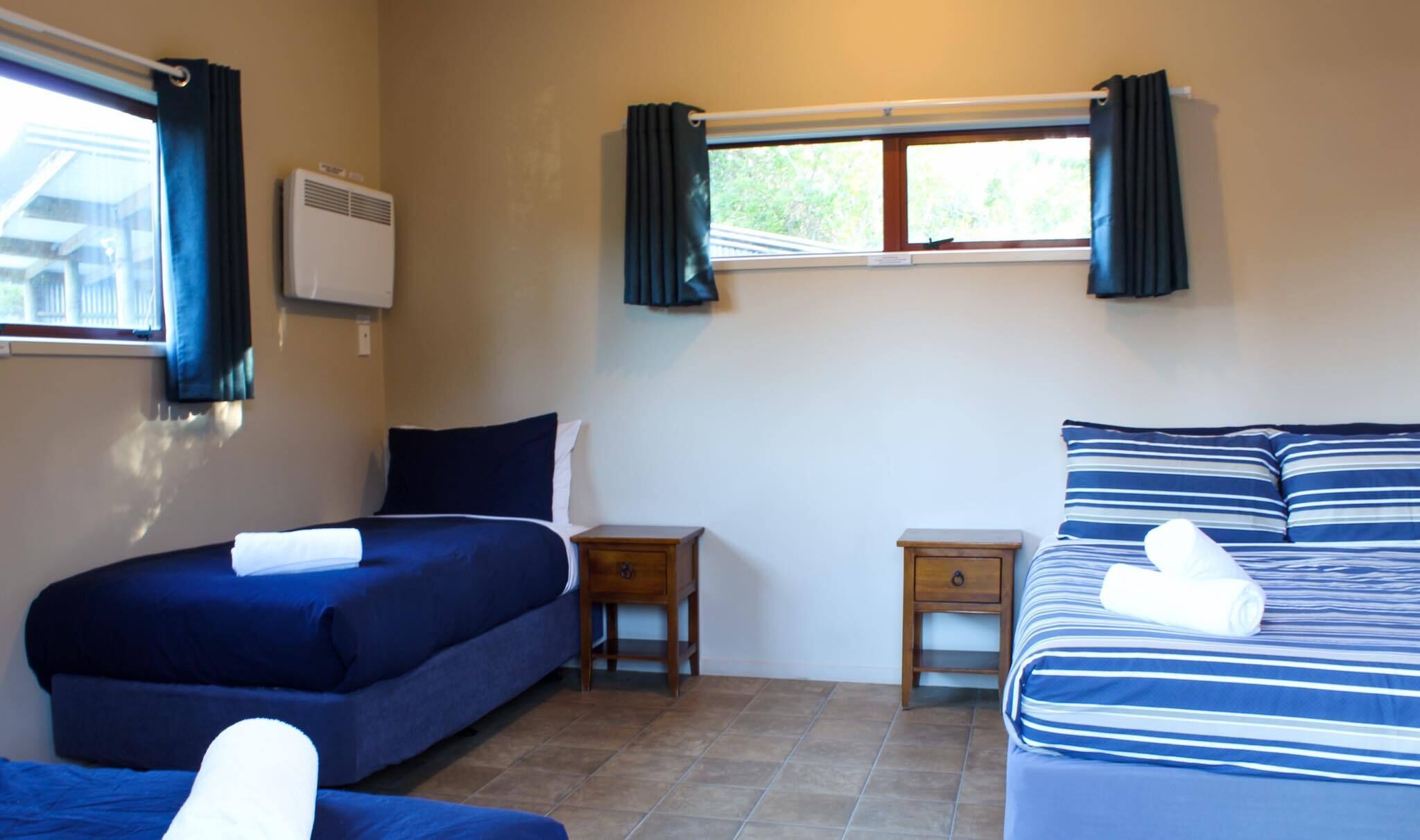 Una habitación con dos camas, una mesita de noche de madera, una ventana con cortinas y un equipo de aire acondicionado.