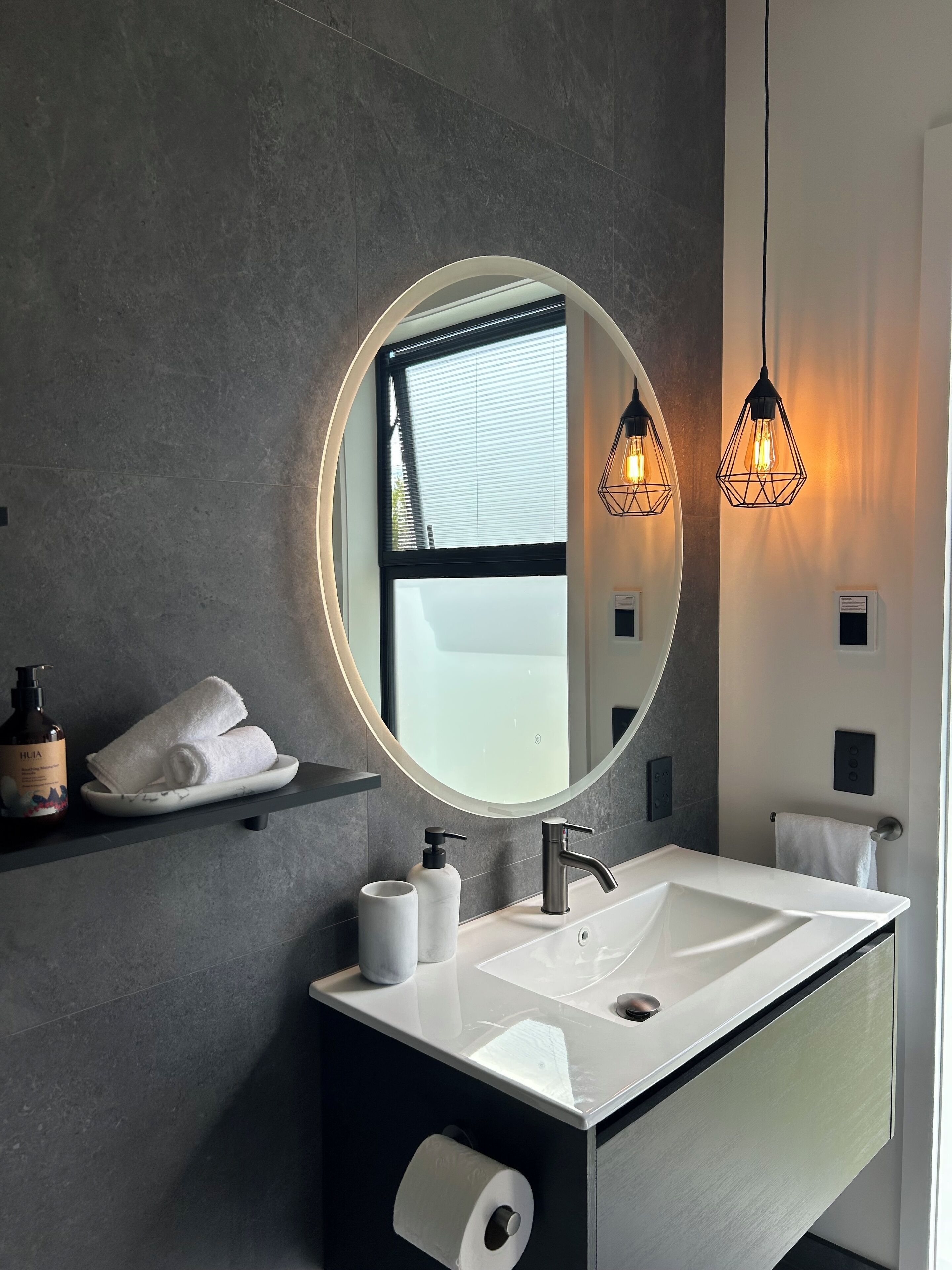 Un baño moderno con un espejo redondo grande, un lavabo blanco y dos lámparas colgantes.