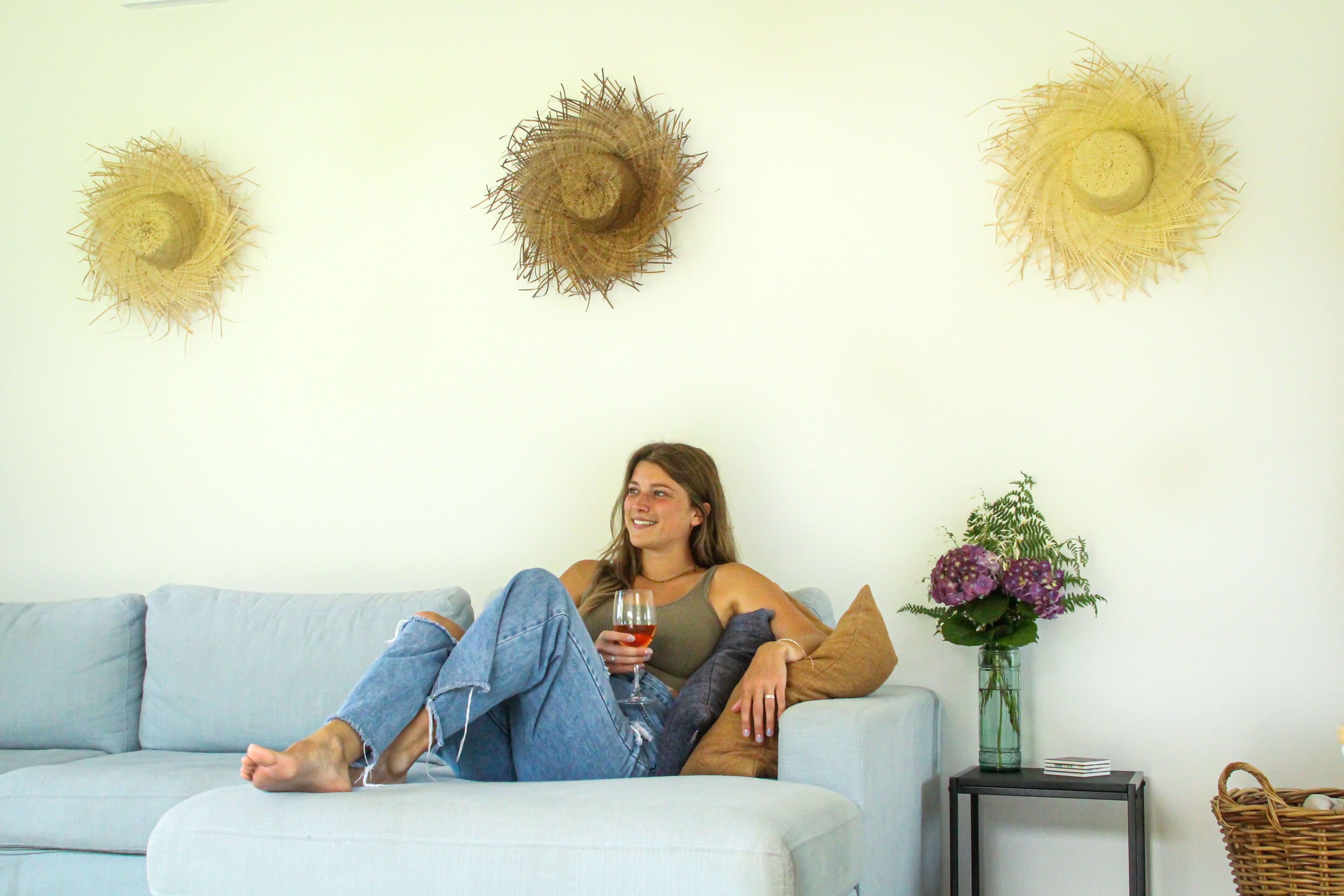 A person sitting on a light blue sofa with a glass of drink, a vase with flowers, and three wall-mounted hats.
