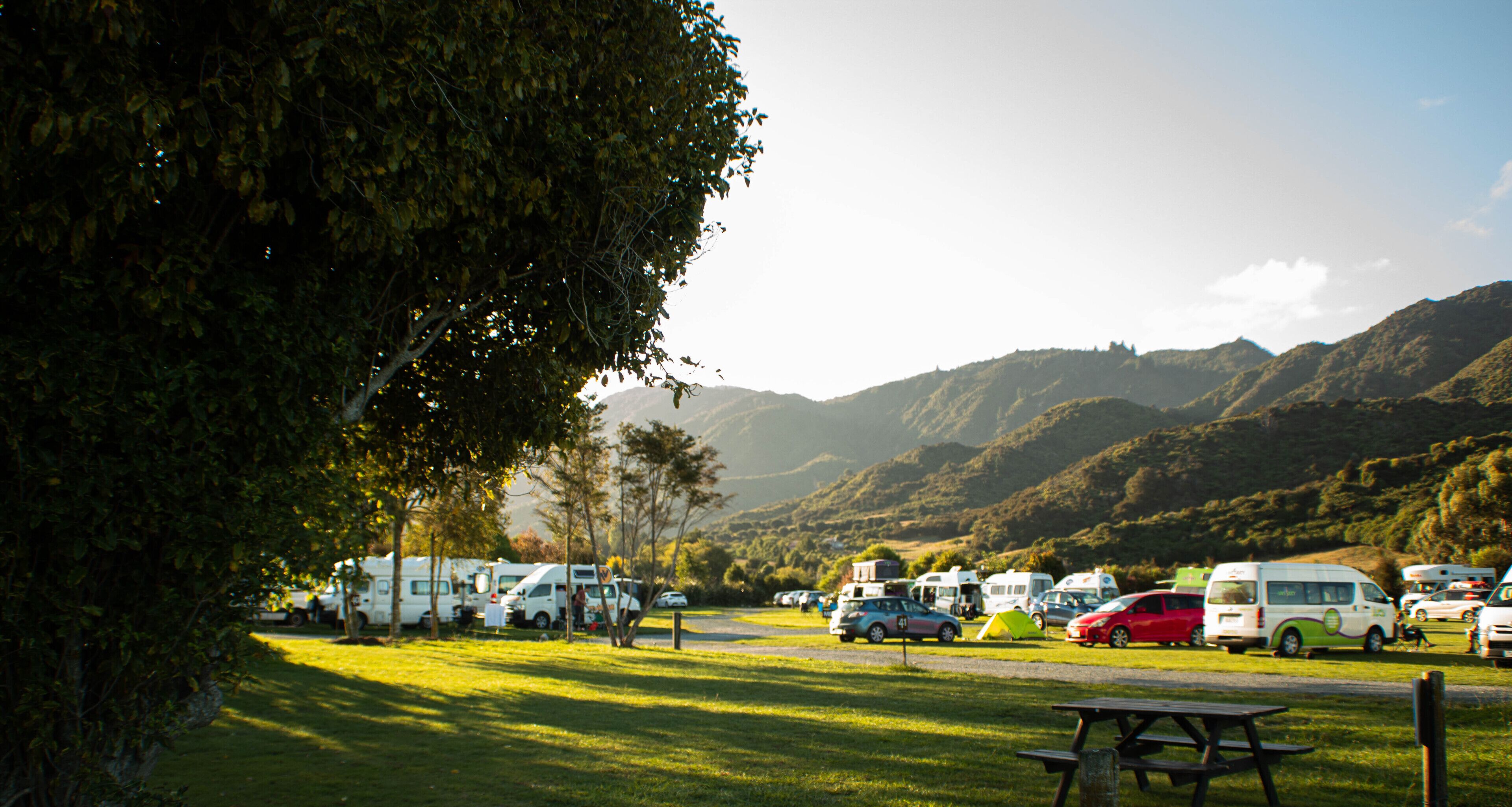 Un camping con vehículos recreativos y automóviles estacionados en un área verde, rodeado de árboles y colinas.