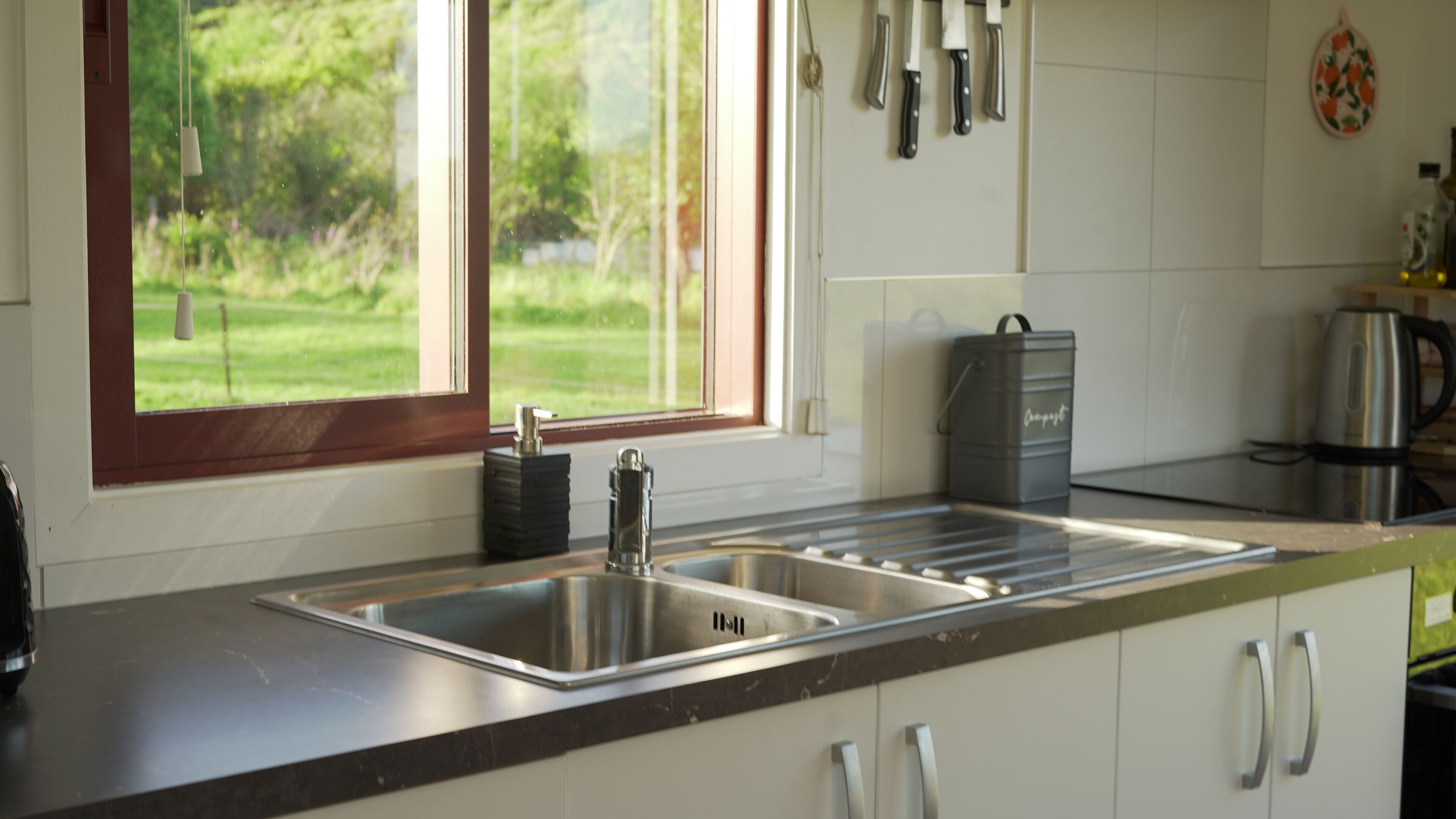 A kitchen with a stainless steel sink, a window with a view of greenery, and a kettle on the counter.