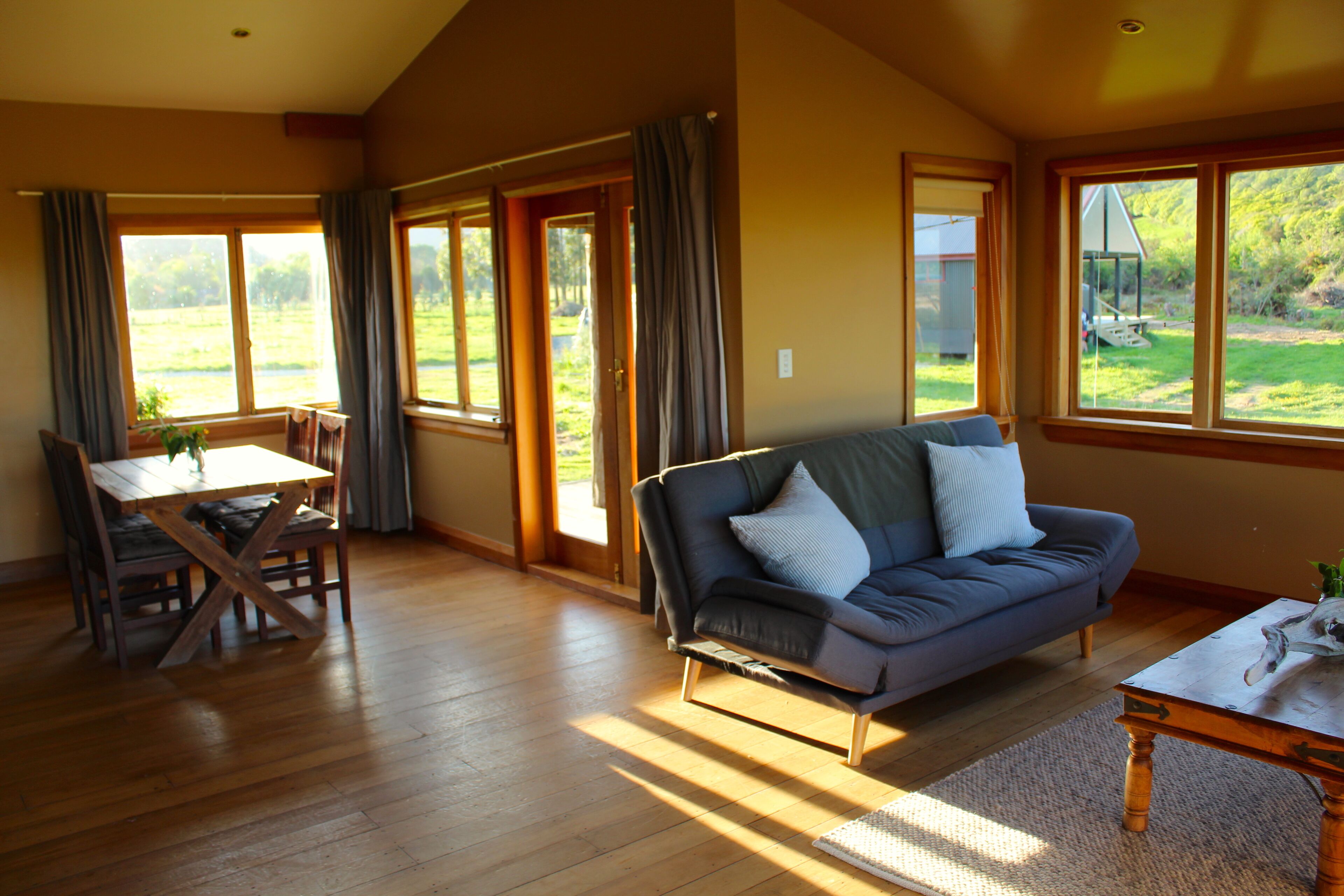 A spacious living area with a wooden floor, a sofa, a dining table, and large windows offering views of a garden.