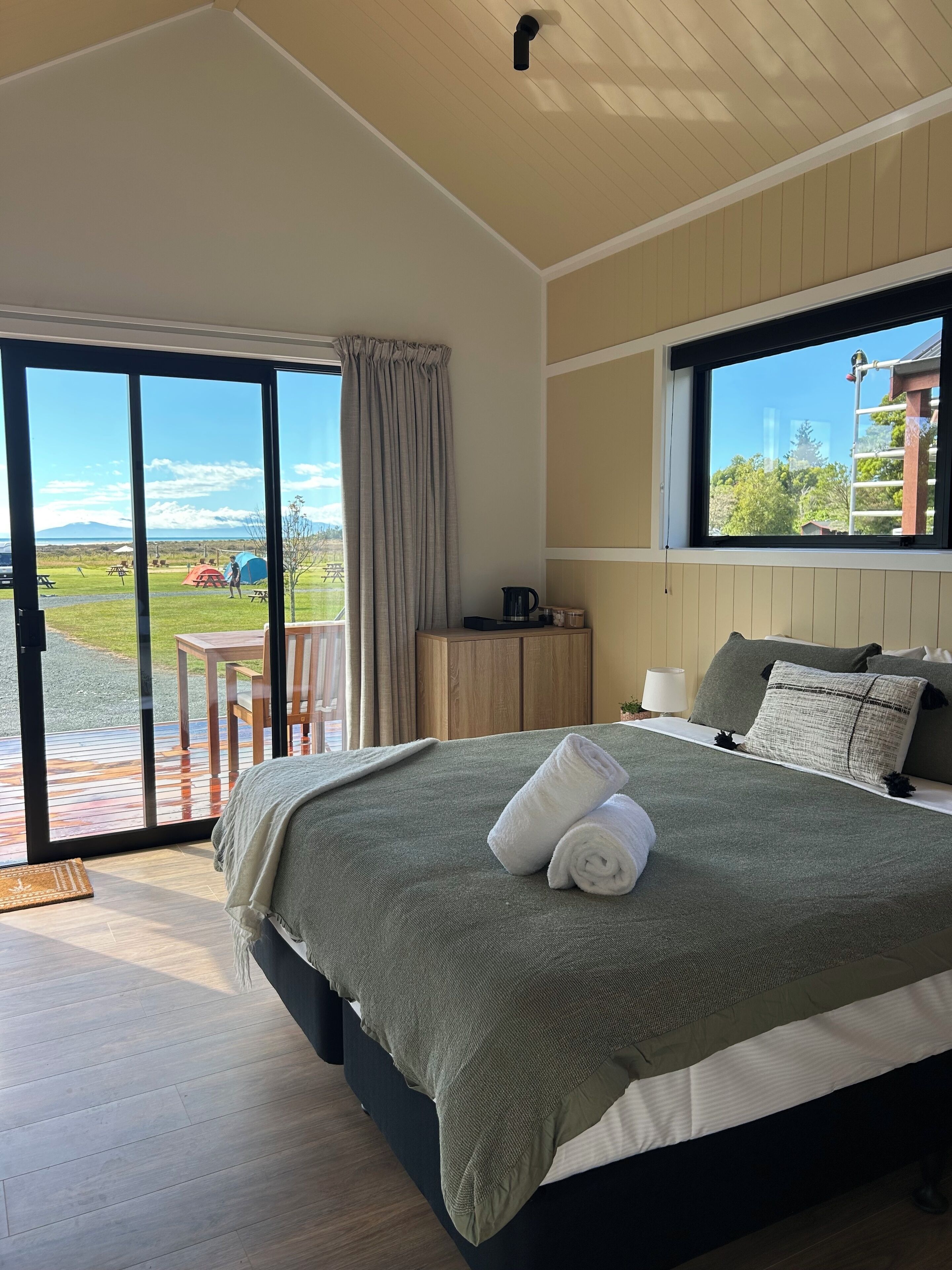 A modern bedroom with a large bed, a view of a grassy area with picnic tables, and a wooden ceiling.