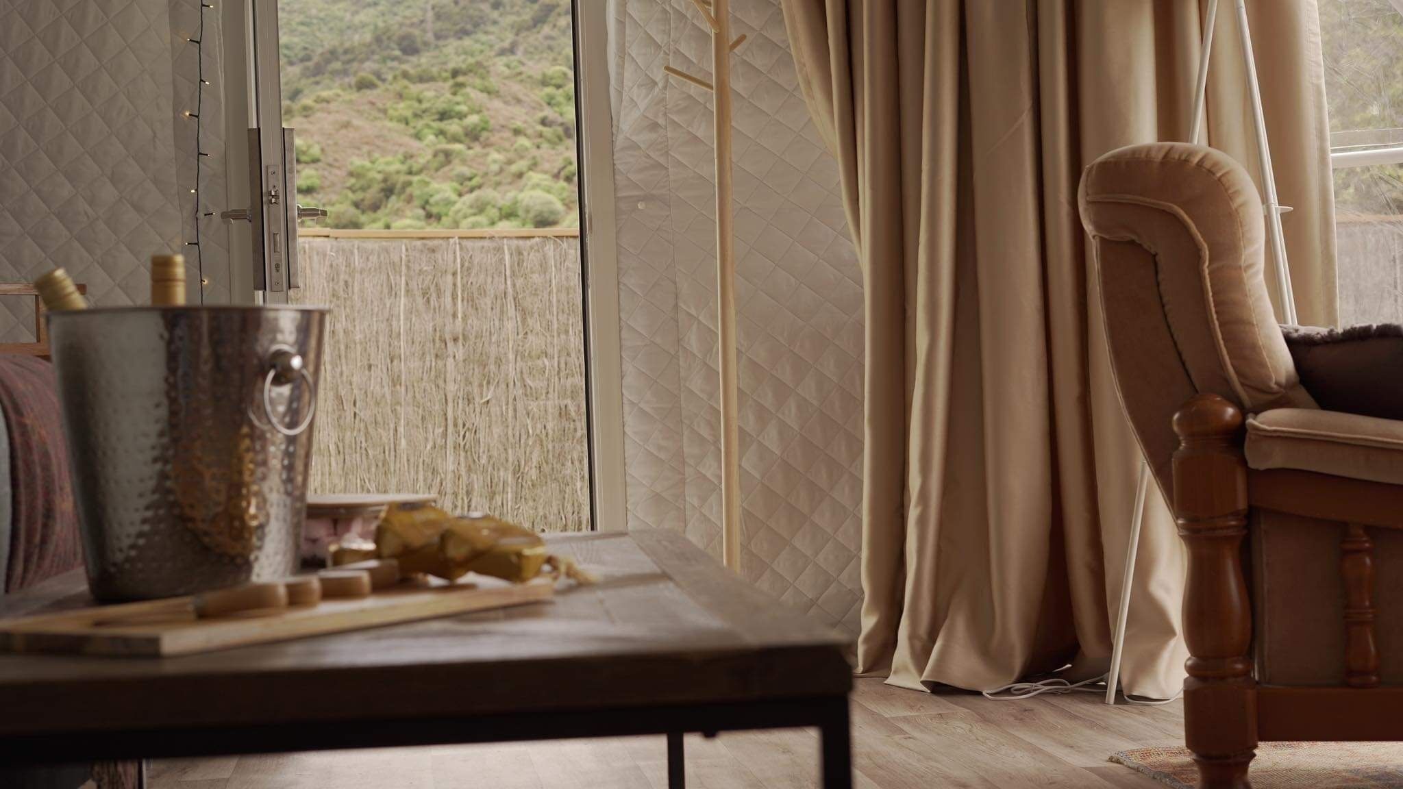 Una habitación con vista a un paisaje verde a través de una ventana, una silla de madera, una mesa con un recipiente metálico y utensilios de madera, y cortinas color beige.