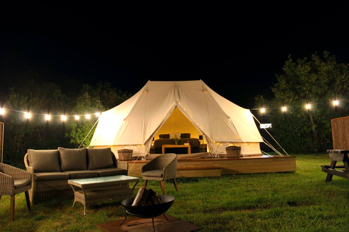 A tent with a flat roof and a central table, surrounded by wicker furniture and a fire pit.