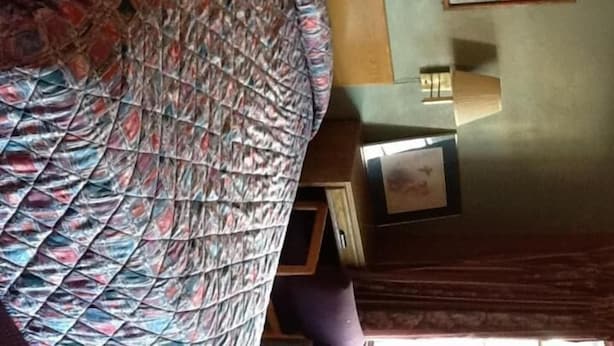 A room with a patterned bedspread, a wooden nightstand with a lamp, and a framed picture on the wall.
