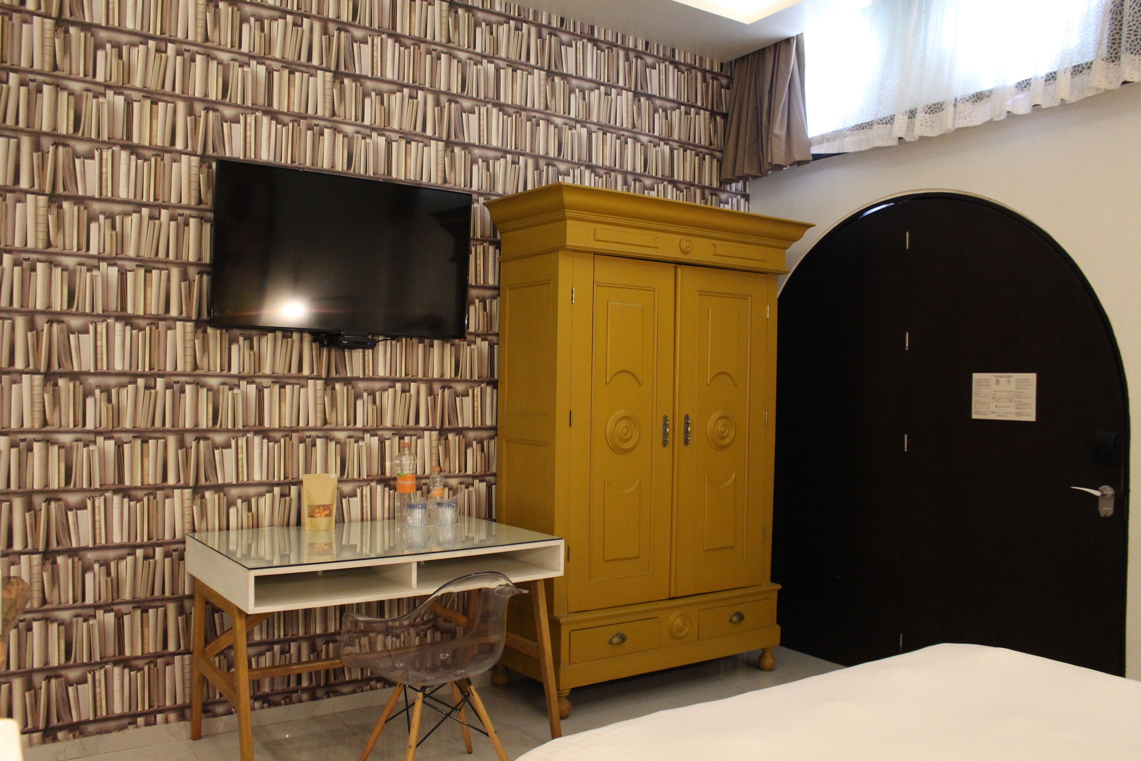 A room with a bookshelf wall, a TV mounted above a glass table, a wooden wardrobe, and a black door.