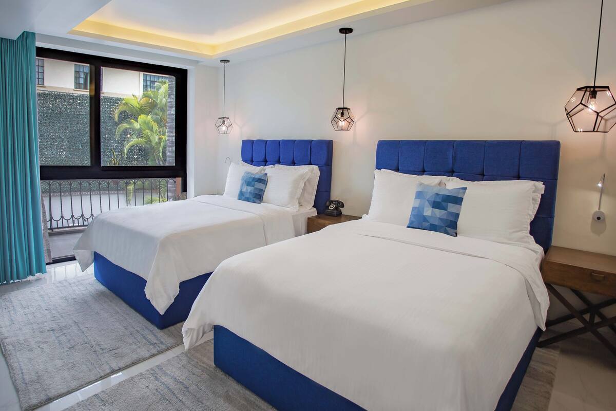 A hotel room with two beds, a large window with a view of greenery, and pendant lighting.