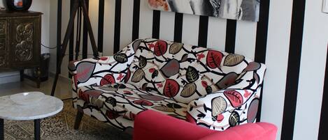 A living room with a red armchair, a patterned sofa, a floor lamp, a small table, and a large abstract painting on the wall.