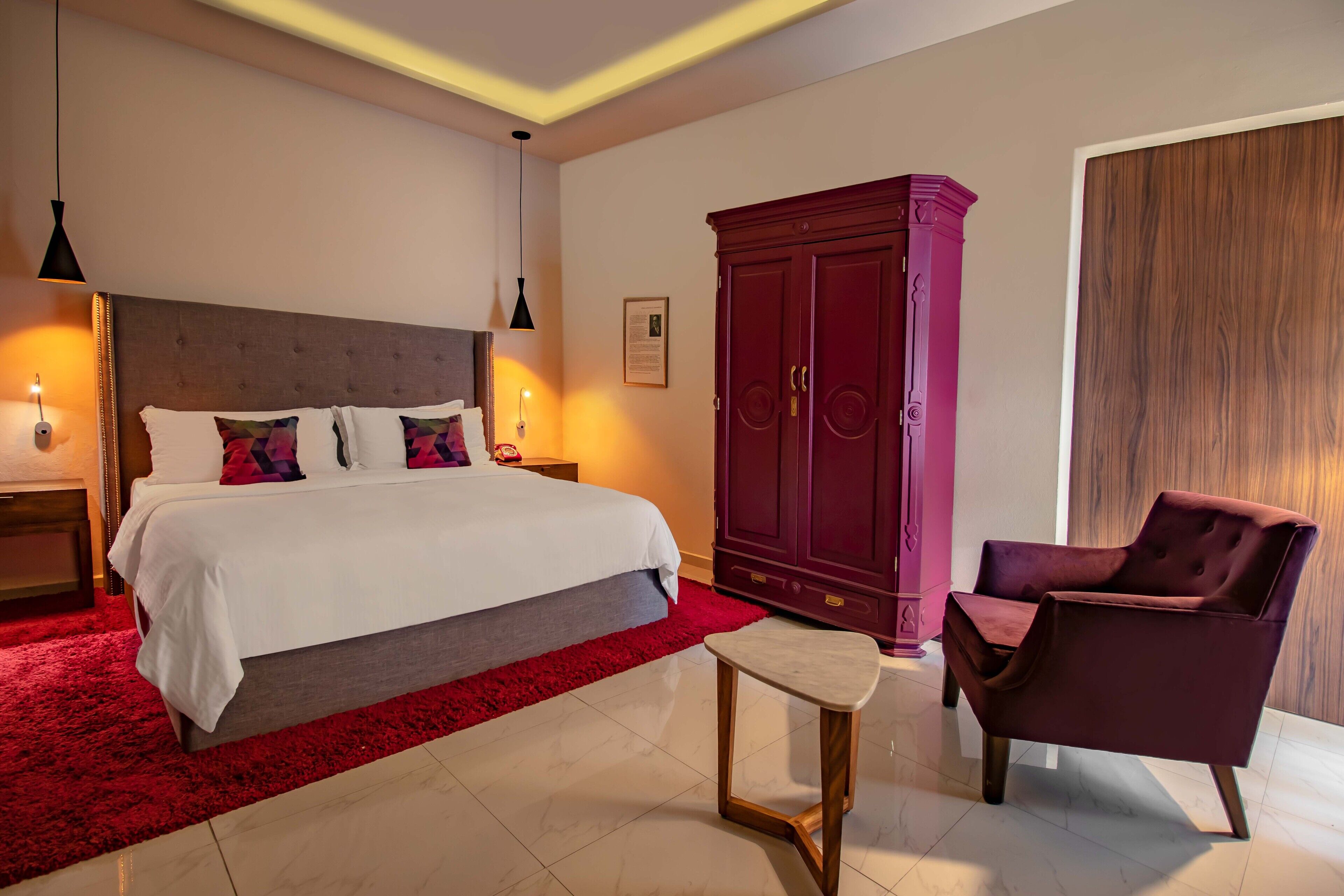 A hotel room with a large bed, a red carpet, a wooden nightstand, a red wardrobe, and a purple armchair.