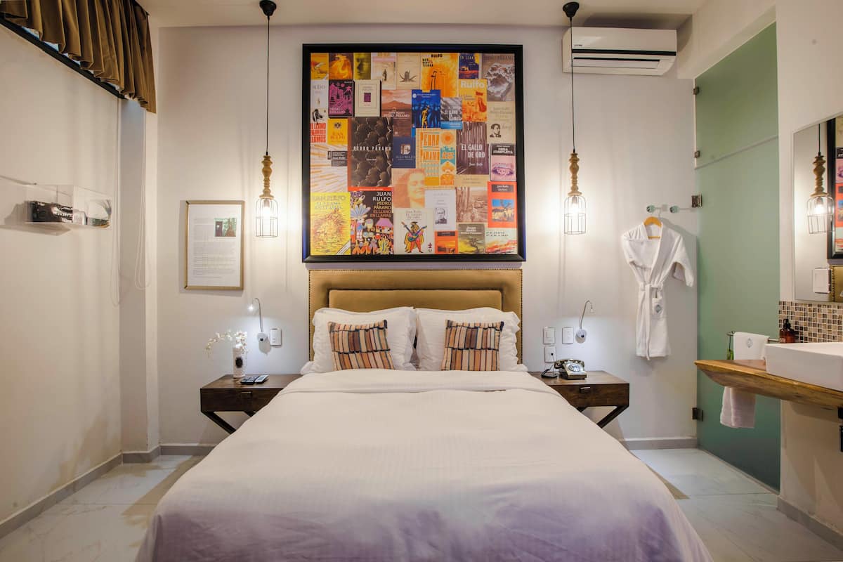 A neatly arranged hotel room with a large bed, bedside tables, a wall-mounted air conditioner, a bathroom with a mirror, and a wall art collage.