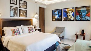 A modern bedroom with a large bed, a glass coffee table, and a chair. There are framed pictures on the wall and a ceiling light.