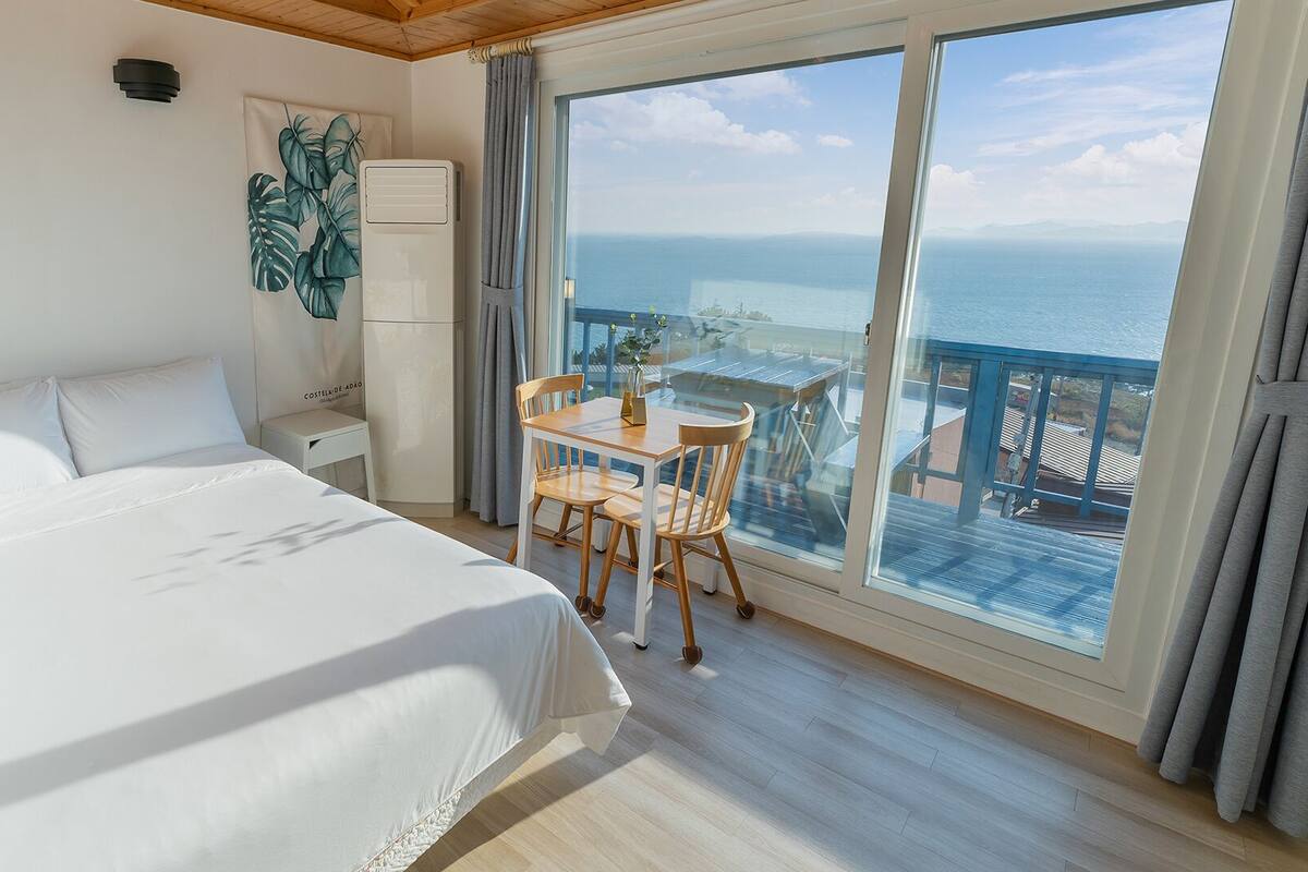 A hotel room with a large bed, a small table with chairs, a nightstand, a wall-mounted air conditioner, and a view of the sea through large windows.