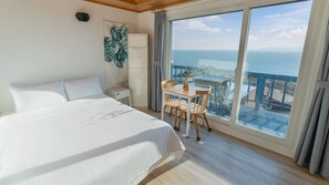A hotel room with a large bed, a small table with chairs, a nightstand, a wall-mounted air conditioner, and a view of the sea through large windows. - Bluegrad Pension (Namhae)