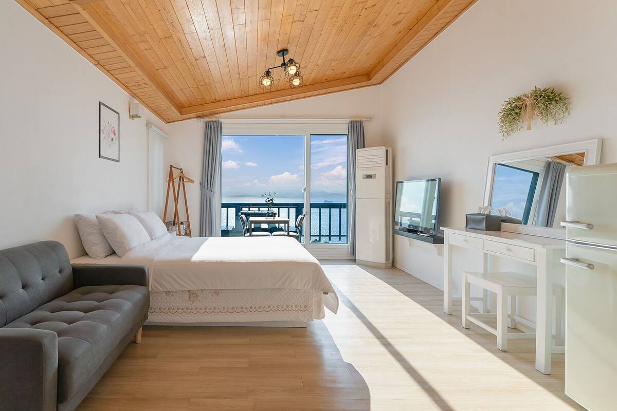 A bedroom with a bed, a sofa, a desk with a TV, and a balcony with a view of the sea.