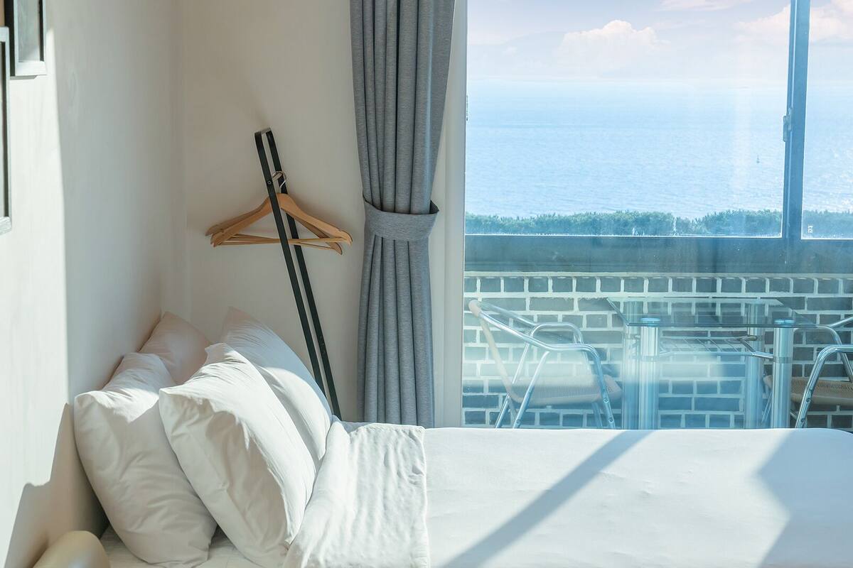 A bedroom with a bed, pillows, a hanging coat rack, and a balcony with a view of the sea.