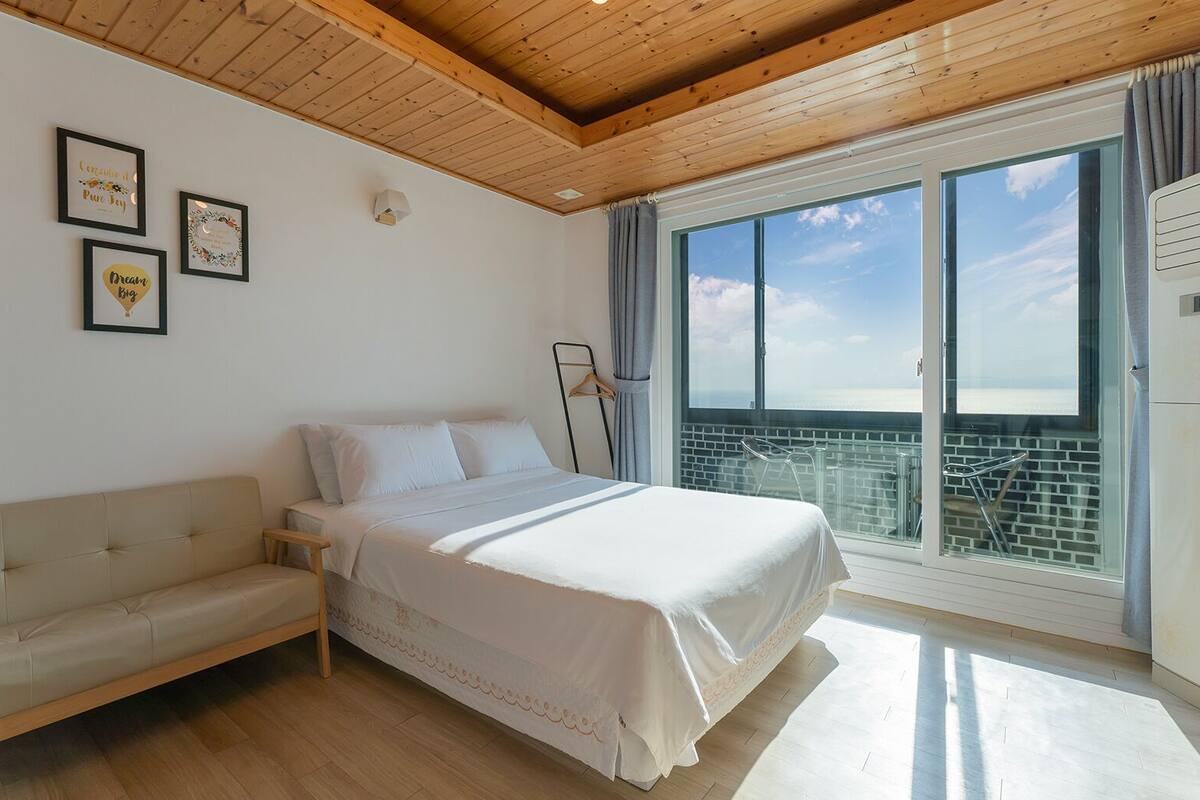 A bedroom with a bed, a sofa, a window with a view, and framed pictures on the wall.