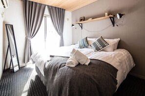 Una cama bien tendida con una colcha blanca y una manta gris, una mesita de noche con una lámpara, y una ventana con cortinas grises.