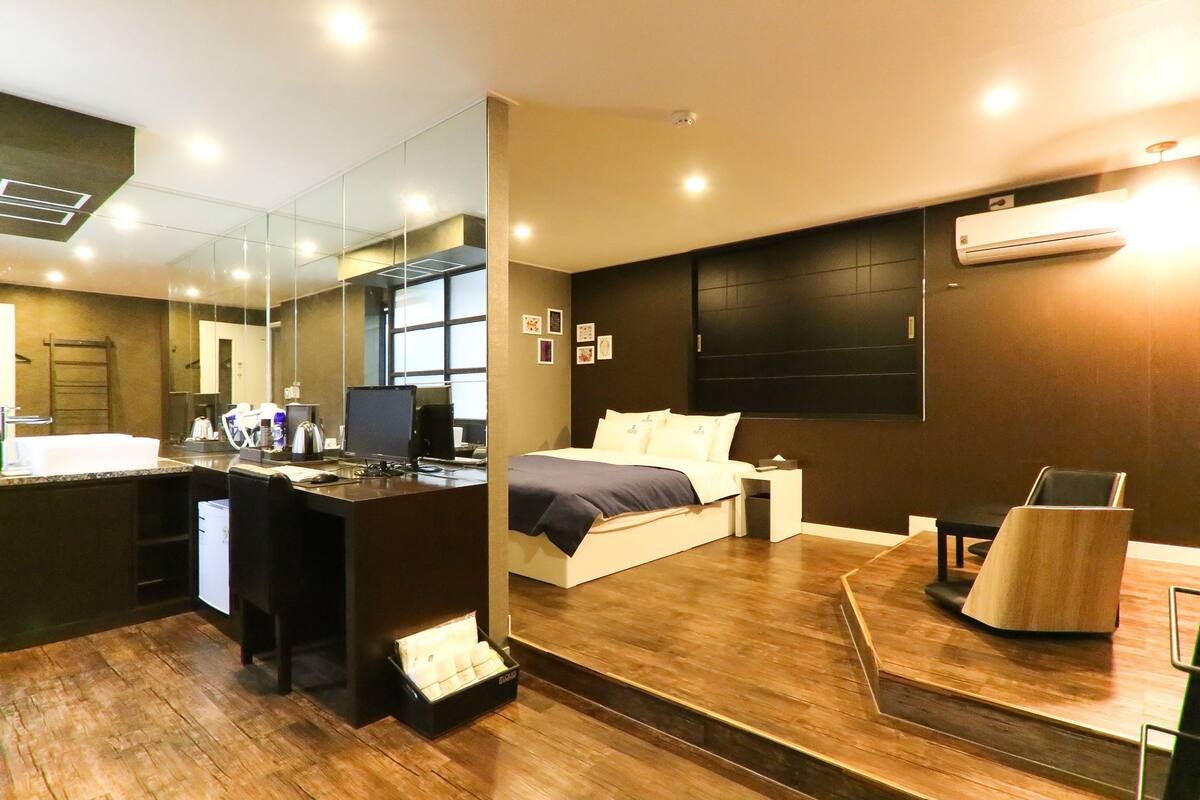 A modern hotel room with a large bed, a desk with a computer, a mirror, and a wooden floor.