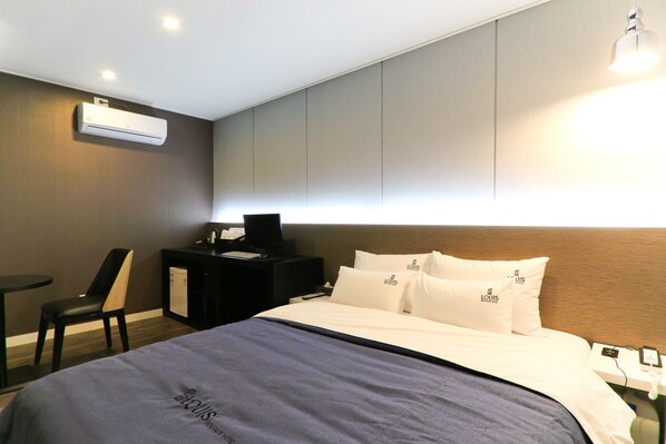 A hotel room with a bed, a desk, a chair, an air conditioner, and a bedside table with a lamp.