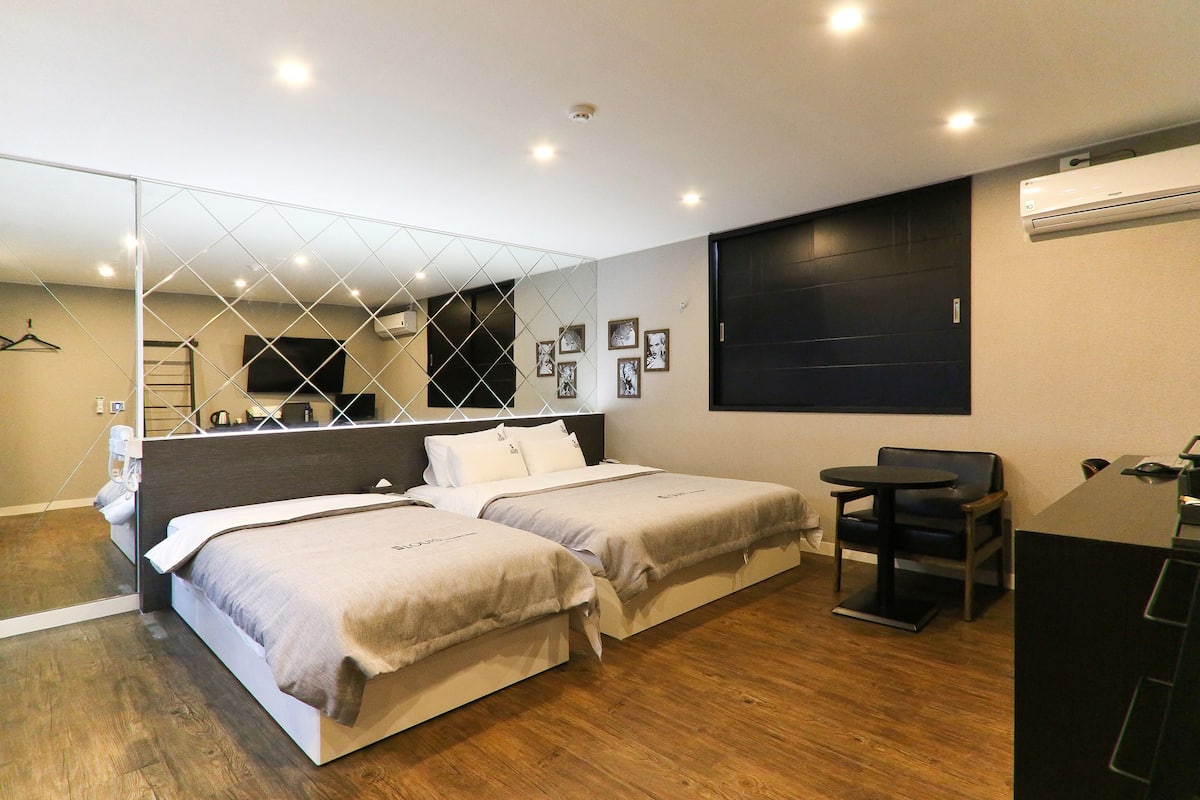 A modern hotel room with two beds, a desk, a chair, a mirror, and a large window.