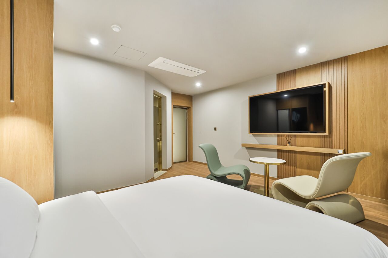 Habitación de hotel con cama, dos sillas, una mesita pequeña, televisor montado en la pared y paneles de madera.