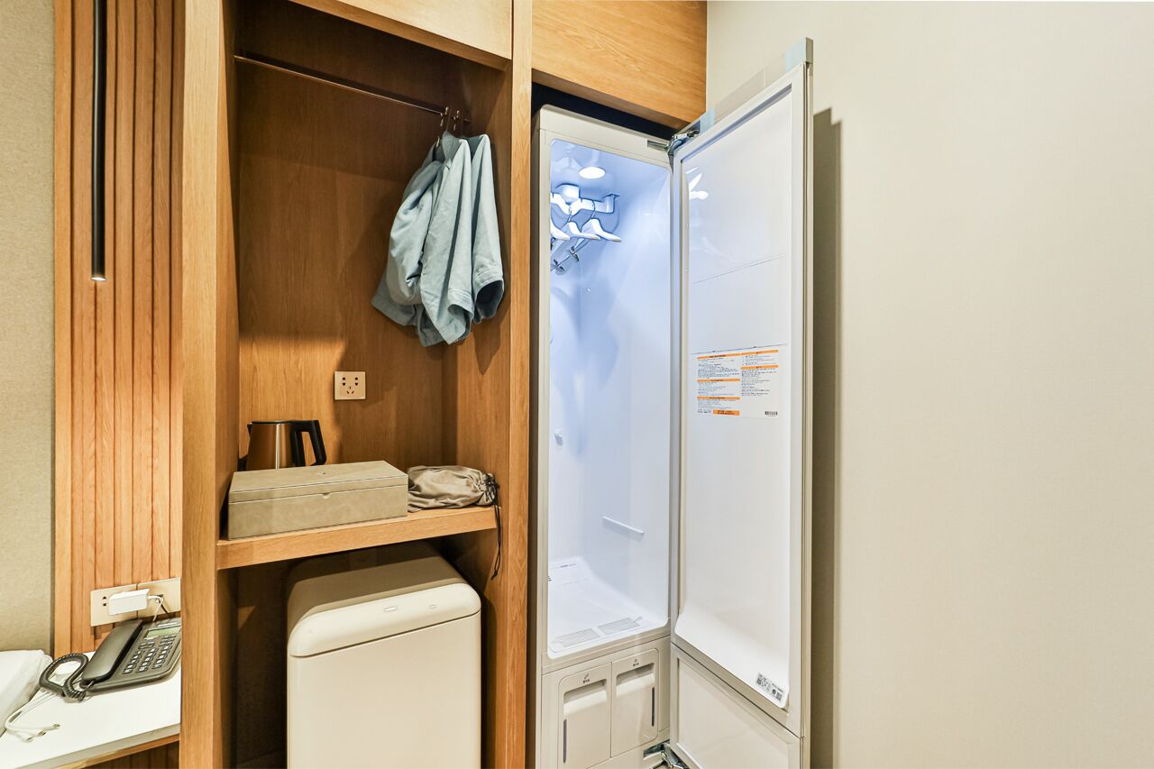 A compact hotel room with a wooden wardrobe, a small desk with a telephone, a compact refrigerator, and a glass shower enclosure.