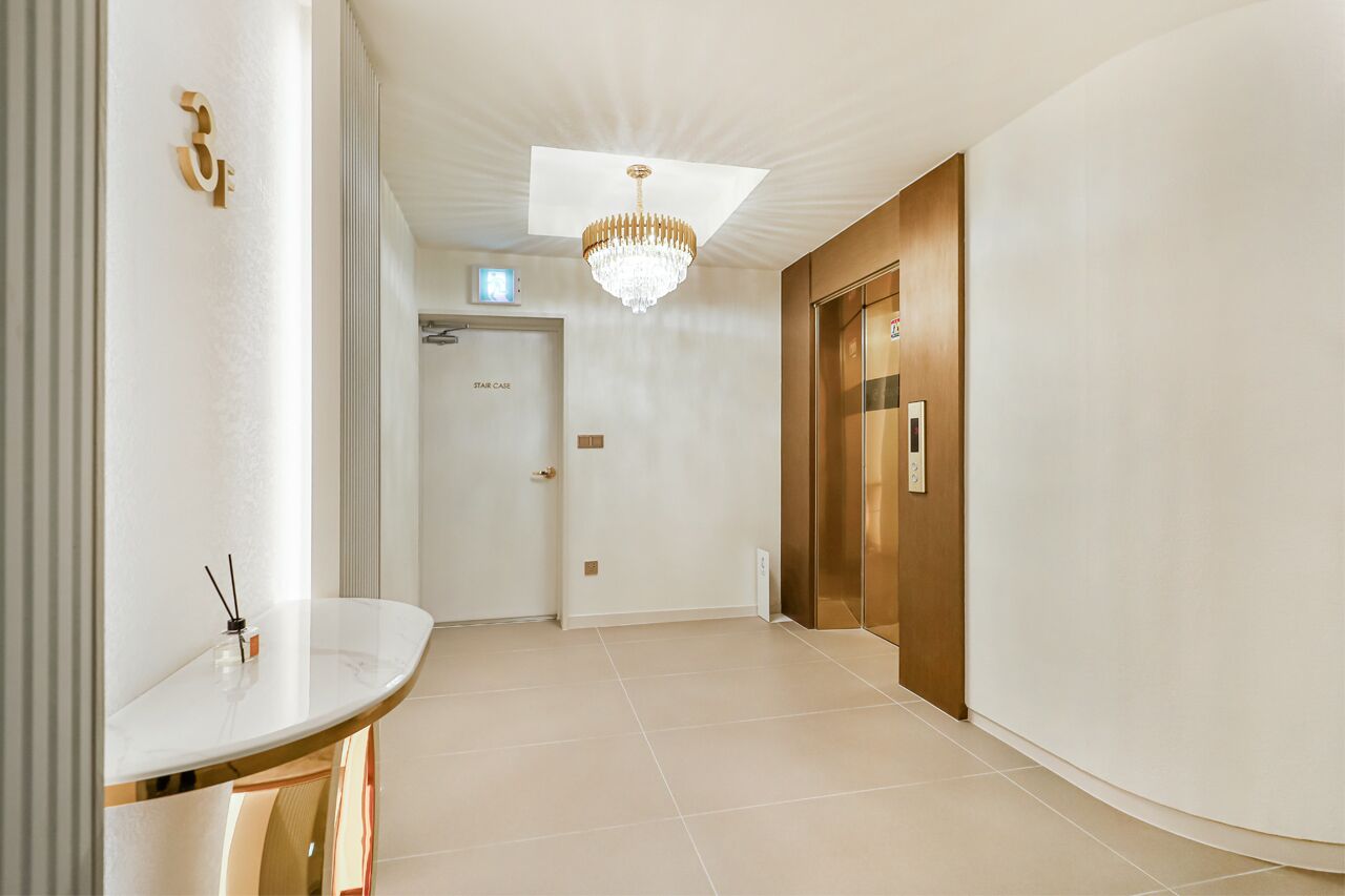 Corredor moderno de hotel com parede branca curva, placa dourada com número, lustre, porta de madeira e um letreiro com a inscrição 'Stair Case'.