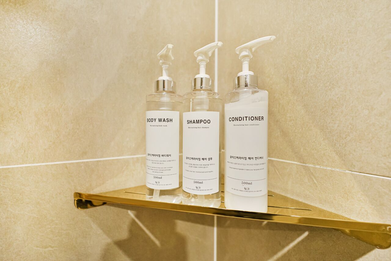 Trois bouteilles de gel douche, de shampoing et d’après-shampoing sont posées sur une étagère dorée, devant un mur recouvert de carreaux.