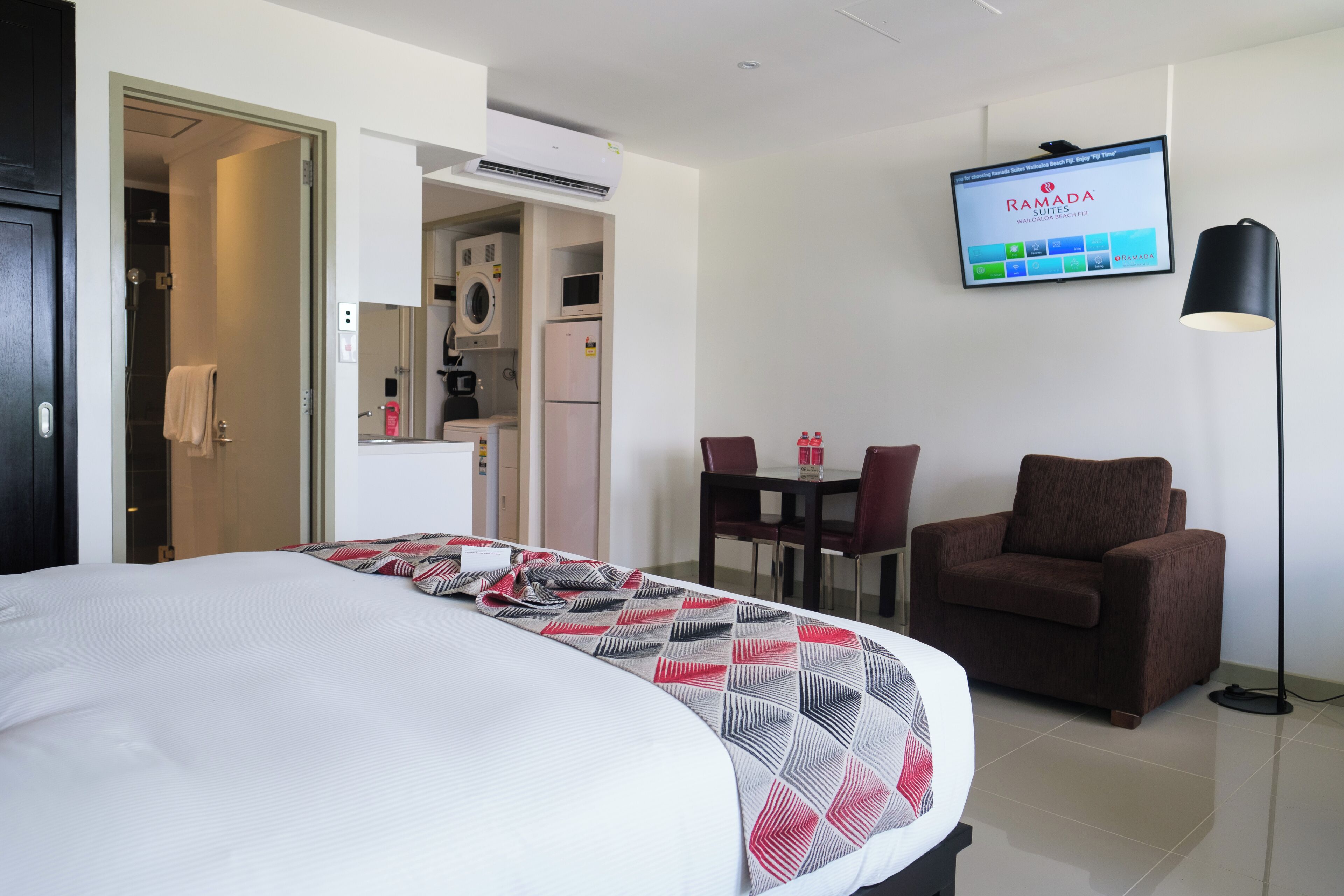 Una habitación de hotel con cama, televisor montado en la pared, mesa de comedor con sillas y un sofá.