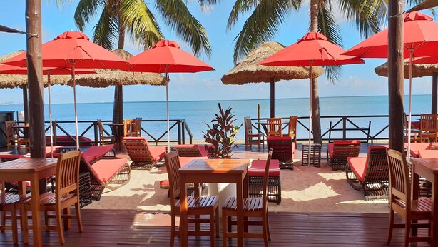 Ein Restaurant direkt am Strand mit Holztischen und -stühlen, roten Sonnenschirmen und Blick auf das Meer.