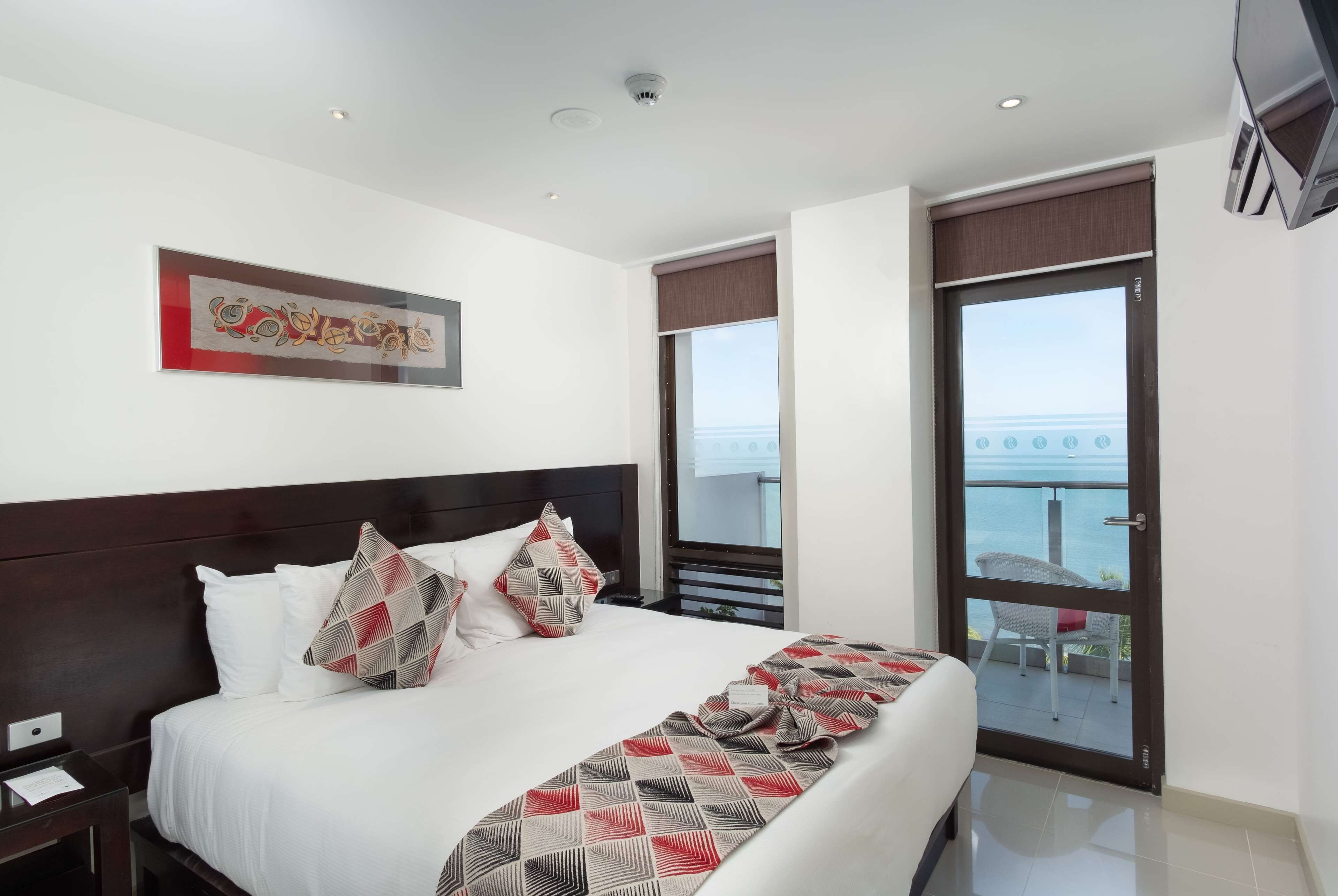 A hotel room with a large bed, a view of the sea through a sliding door, a ceiling with recessed lighting, and a framed artwork on the wall.