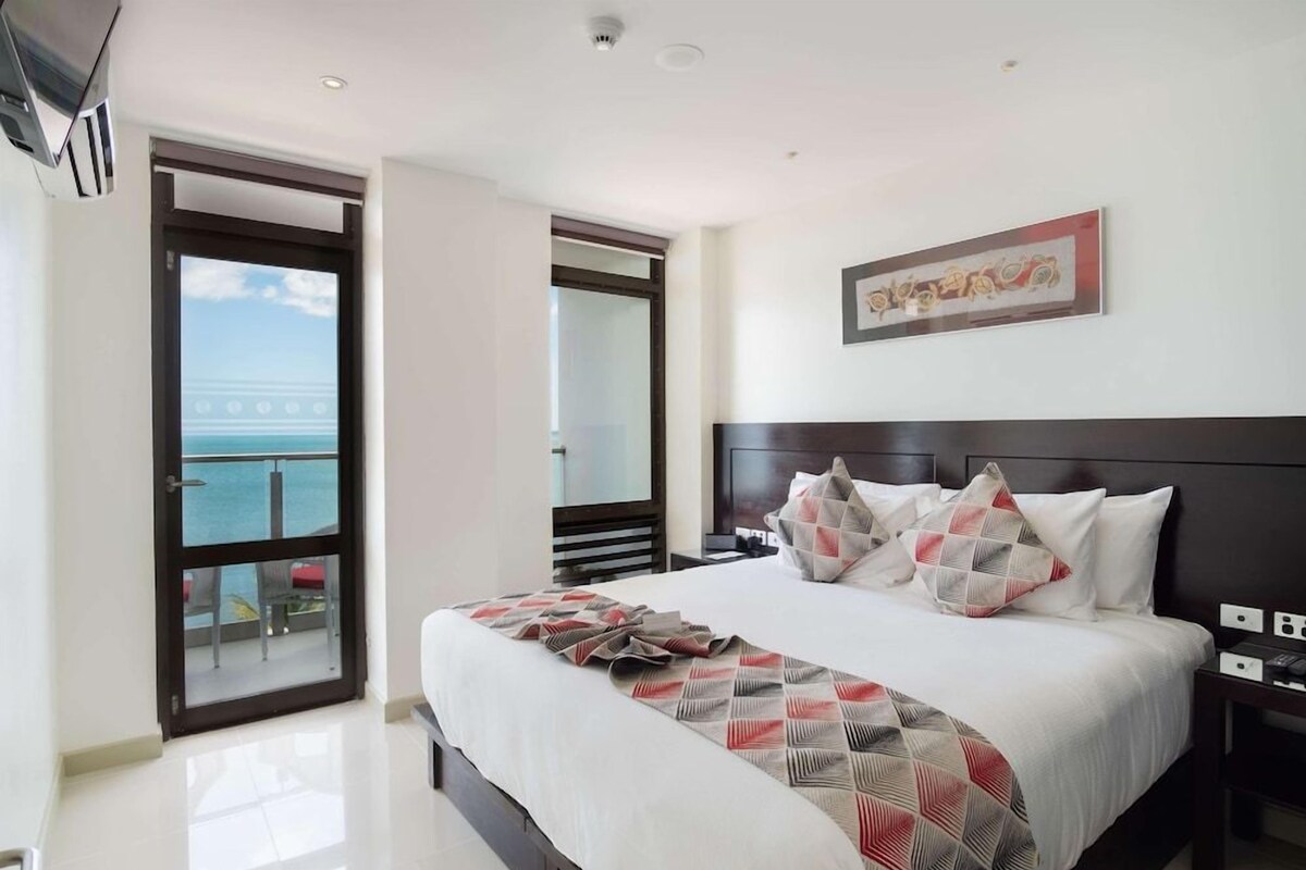 A hotel room with a large bed, a view of the sea through a sliding door, and a painting on the wall.
