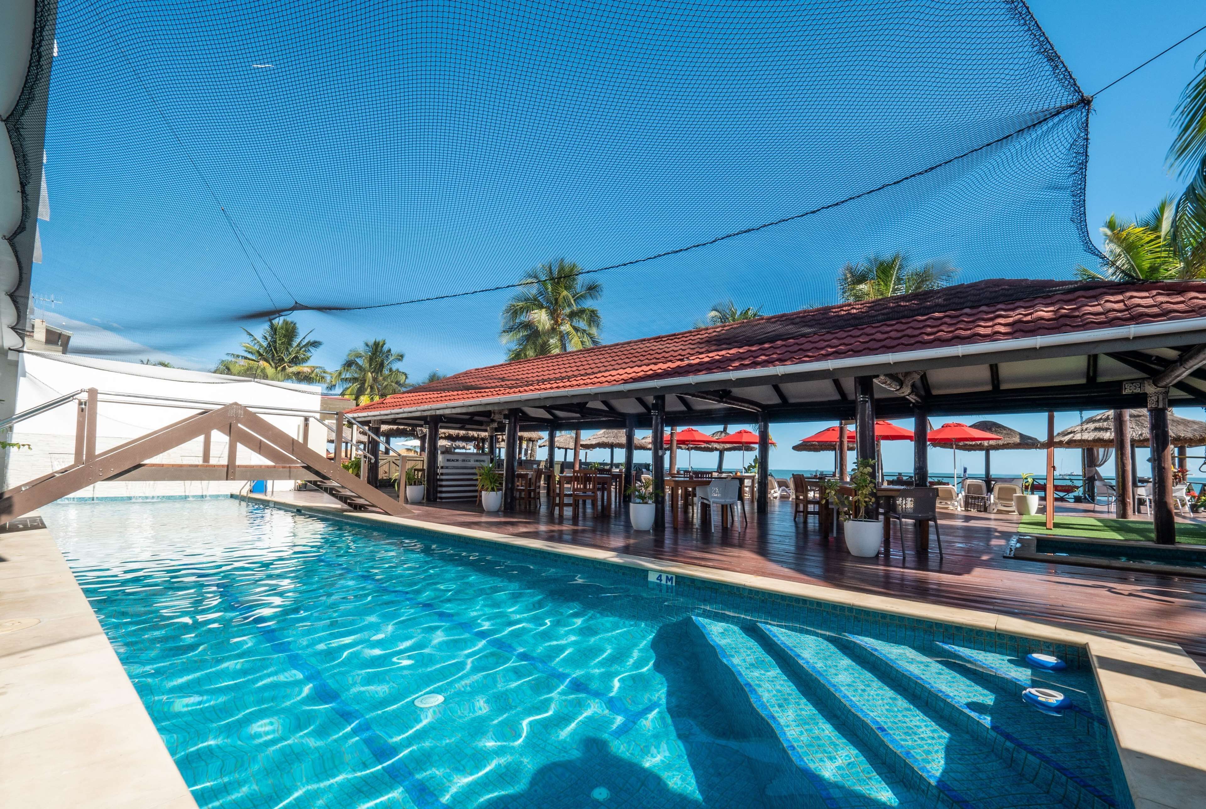 Una piscina amb un solàrium de teulada vermella, palmeres i un cel blau clar.