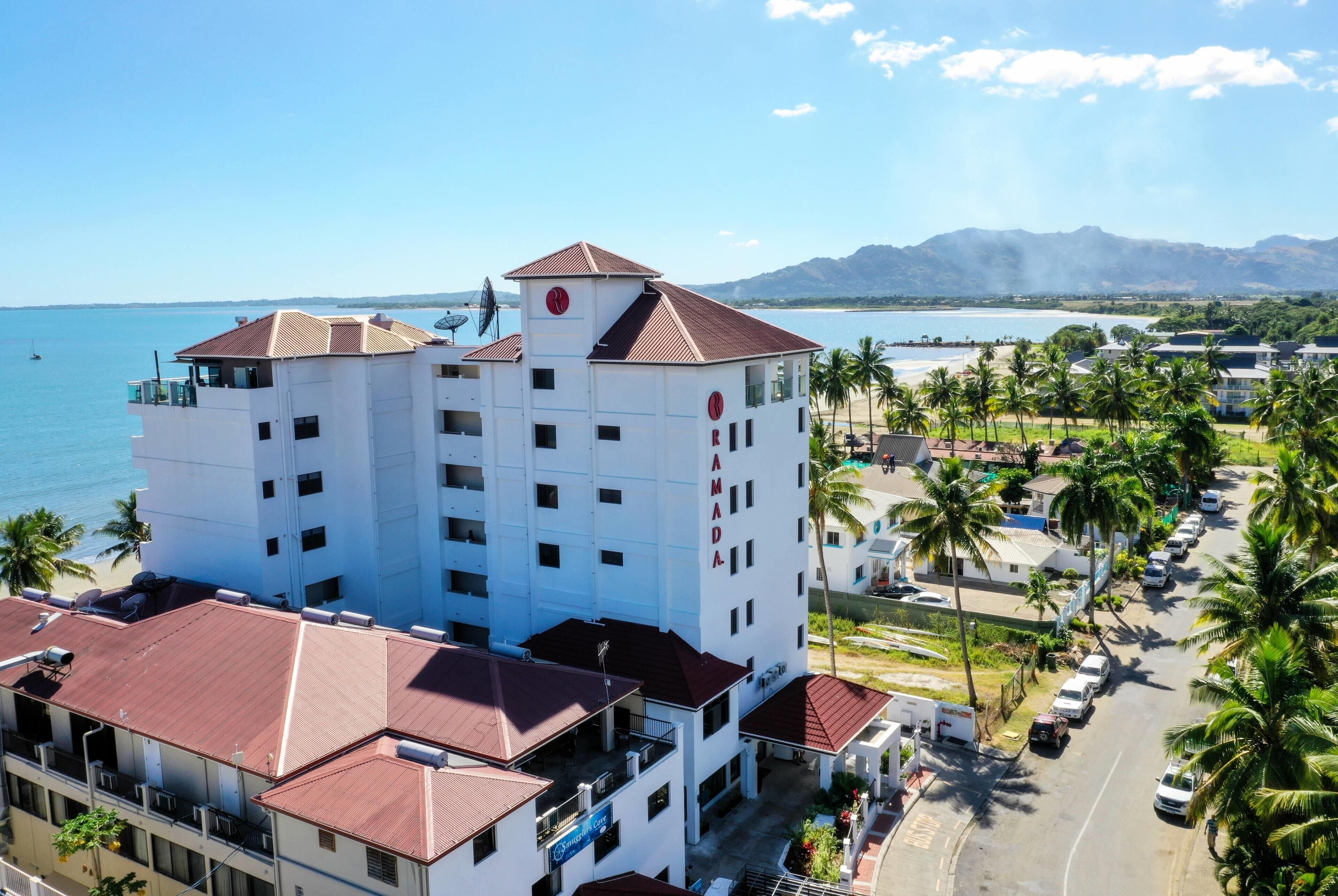 A multi-story hotel with a central tower, a beach view, and a parking area with cars and palm trees.