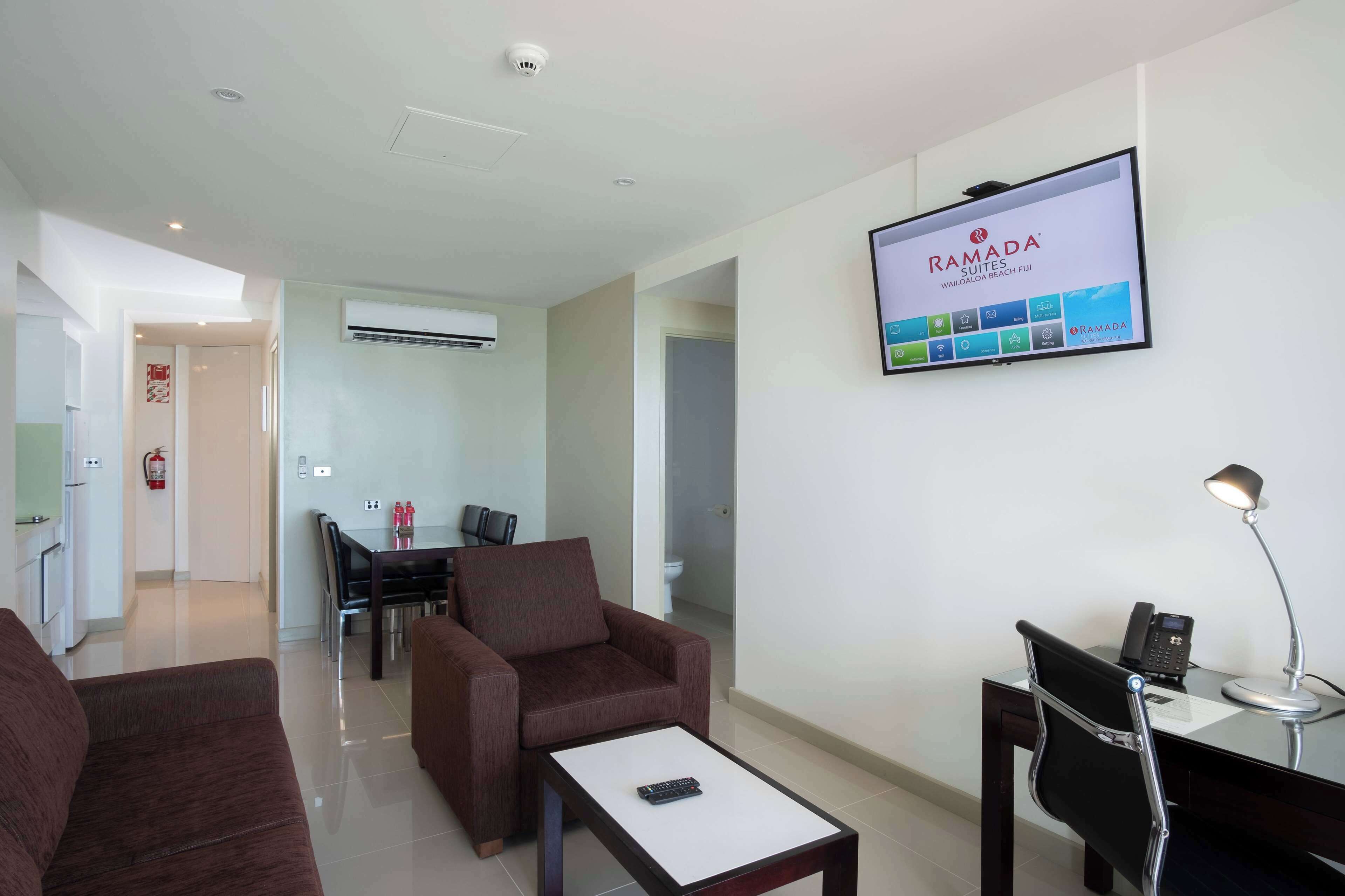 Una habitación de hotel moderna con una televisión de pantalla plana mostrando la marca Ramada, un sofá, una mesa de centro, un escritorio con una lámpara y una silla.